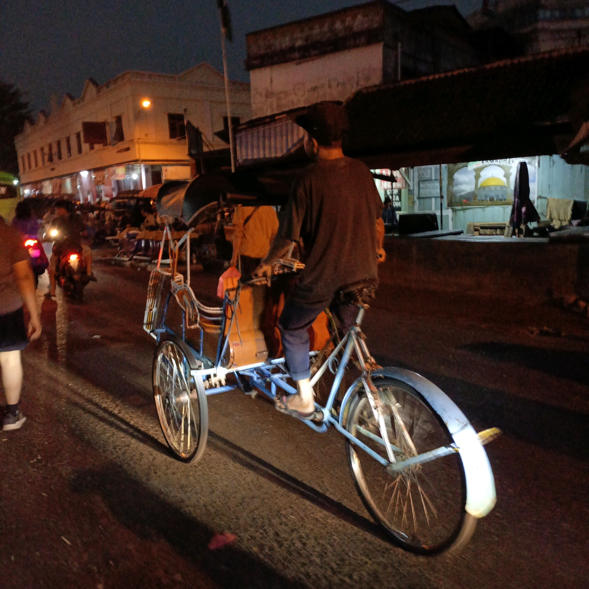 A pedicab driver pedals a light blue pedicab down a street at night in Salatiga, Indonesia. The driver is wearing a dark shirt and blue pants. The pedicab is carrying an orange bag. The street is paved and wet. There are buildings on either side of the street. The buildings are lit by streetlights and the lights from inside. The sky is dark and there are some clouds. A motorcycle is visible in the background.