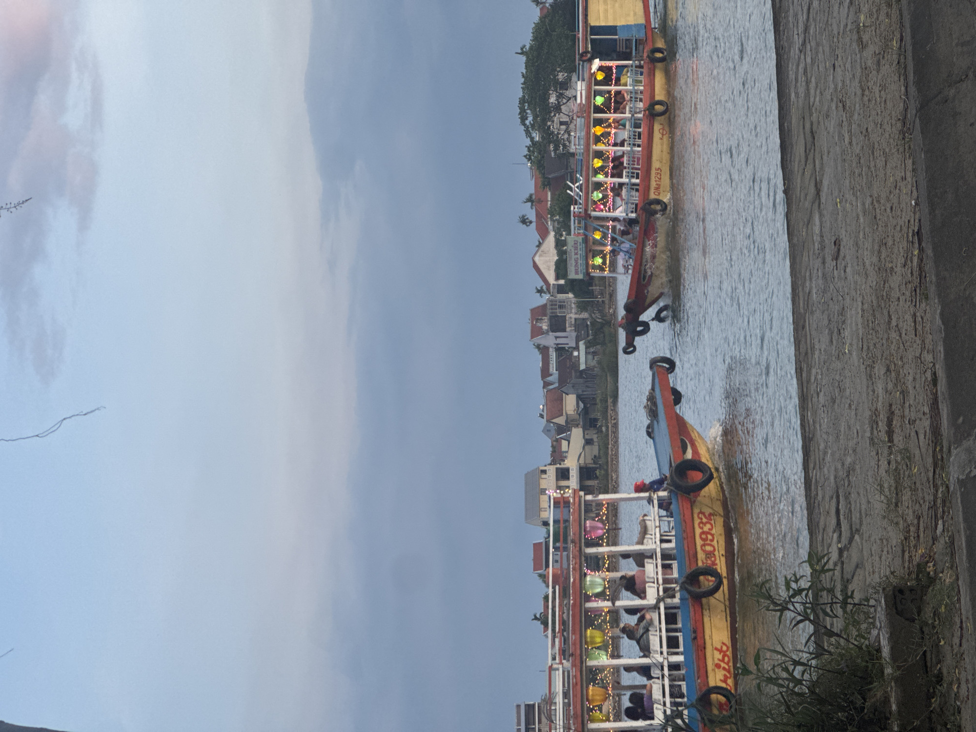 Two boats are sailing on a waterway. One boat has the numbers "90932" displayed. A group of people are present on the boats. They are near the banks of Hội An Ward, Vietnam. The environment is an urban, built-up area.