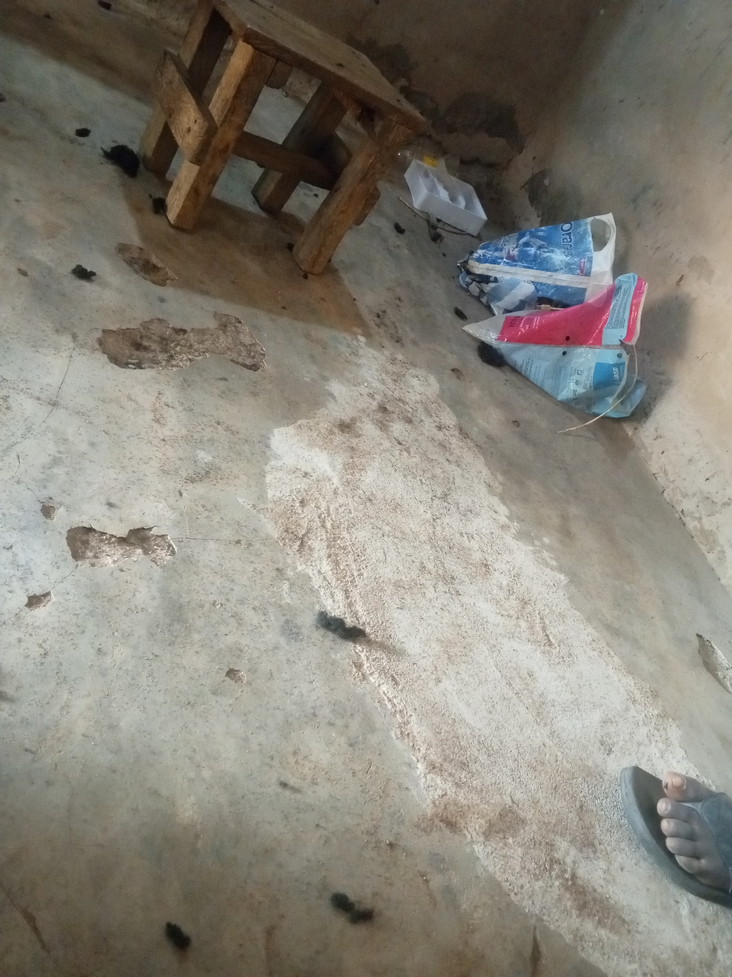 The image depicts a close-up, low-angle view of a concrete floor with a wooden stool and some plastic bags in the background. The floor is visibly dirty and cracked, with patches of discolored concrete and some dark debris scattered across its surface. A wooden stool, constructed from rough-hewn planks, is positioned in the upper left quadrant of the image, casting a shadow on the floor. To the right of the stool, several plastic bags, including a blue and white one with text and a pink and blue one, are piled up. A white styrofoam container is also visible near the stool. In the bottom right corner, a person's foot wearing a dark sandal is partially visible, resting on the floor. The environment appears to be an indoor space, possibly a room or a covered area, with a sense of disarray and neglect. There are no explicit indicators of the specific location within Nyabe, Nigeria, beyond it being an urban or semi-urban setting suggested by the materials and objects present.