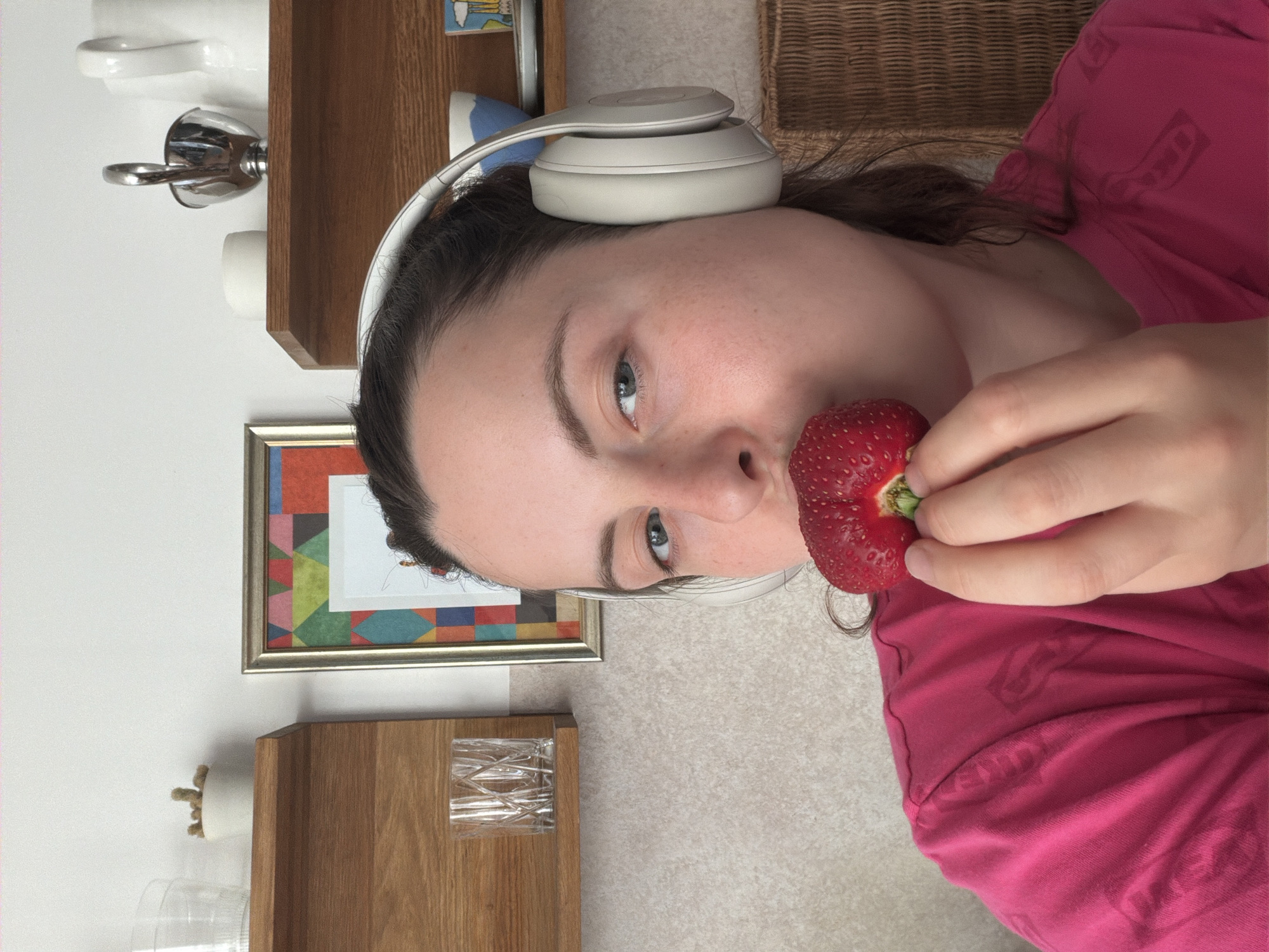 The image shows a person wearing white headphones, lying down with a strawberry held up to their mouth. The person is wearing a bright pink shirt with a repeated pattern of abstract shapes. Their eyes are open and looking towards the camera, with a slight smile. The background includes a wooden shelf with various objects, including a framed picture with a geometric design and a woven basket. The lighting suggests it might be daytime, possibly indoors. The overall mood appears casual and relaxed.