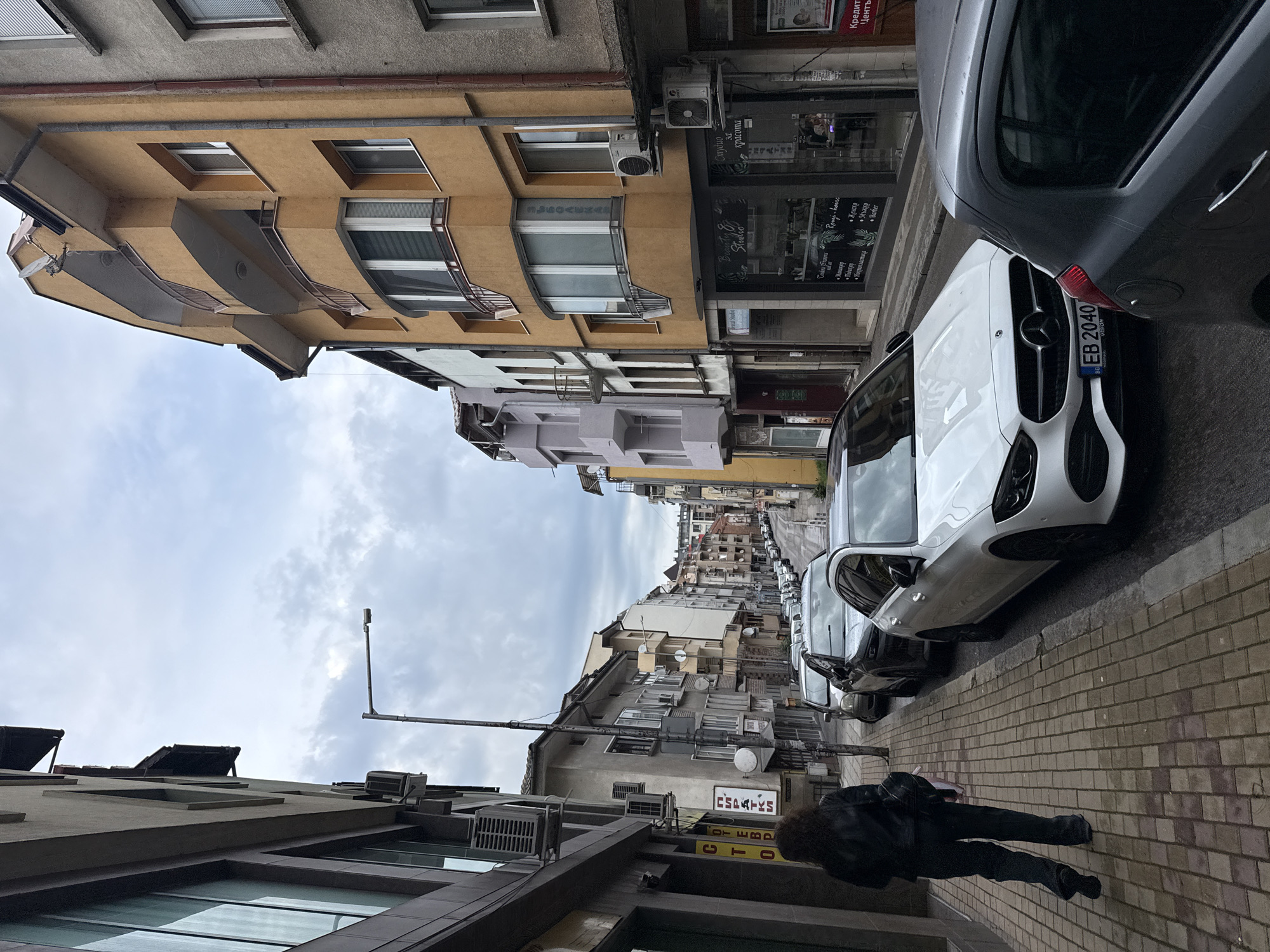 The image depicts a street scene in Gabrovo, Bulgaria. The view is looking down a narrow street flanked by buildings of various heights and architectural styles, predominantly painted in shades of yellow, grey, and beige. Parked cars line the right side of the street, including a white Mercedes with the license plate "EB 2040 VISION". On the left side of the frame, a person dressed in black lies or falls on the sidewalk. Signage in Cyrillic script is visible on the buildings, and some air conditioning units protrude from the facades. The sky is overcast with grey clouds.