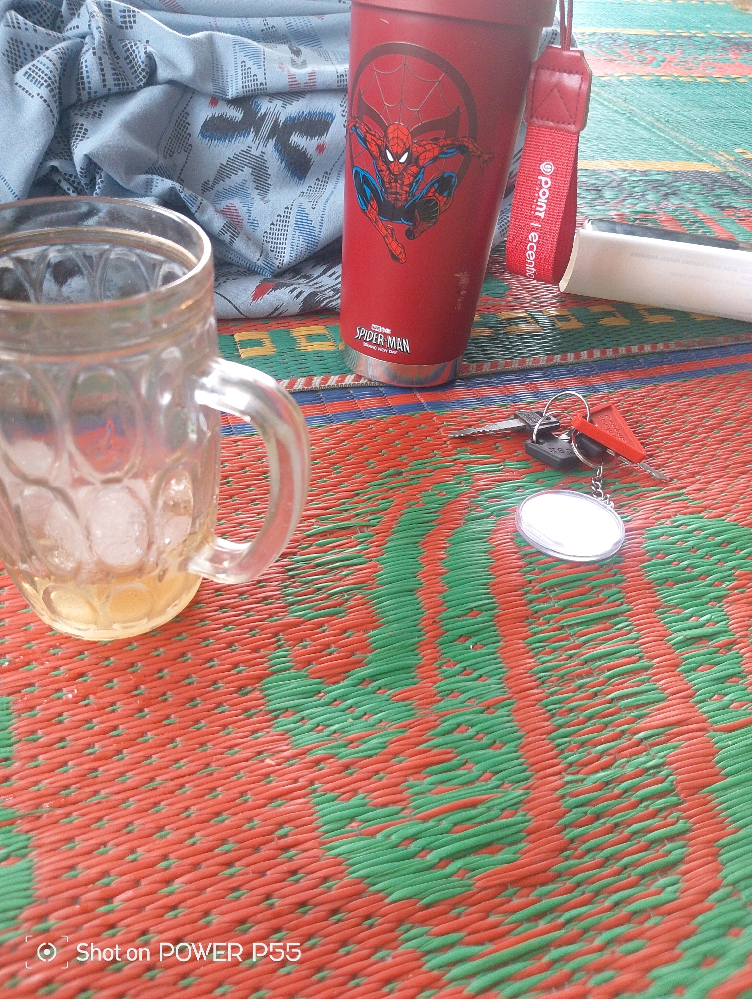 An overhead shot of objects placed on a woven mat in Slawi, Indonesia. To the left, a glass mug filled with ice and a light-colored liquid rests on the mat. Next to it, a red thermos with a Spider-Man graphic is upright. A red strap with white text, "Point", hangs from the thermos. To the right, a set of keys with a red fob and a circular keychain lies on the mat. A white rolled-up object, possibly a book or notepad, is partially visible behind the thermos. The mat is woven with red and green threads. The bottom left corner displays the text "Shot on POWER P55".
