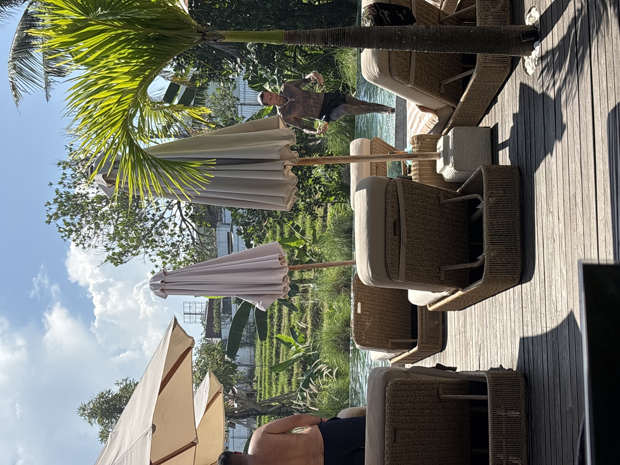 The image shows an outdoor scene in North Kuta, Indonesia, beside a pool. Three wicker lounge chairs with white cushions are positioned on a wooden deck, shaded by large white umbrellas. A palm tree is situated beside the pool. A man with short, light hair is partially visible, reclining in one of the chairs. Another man is seen mid-air above the pool. Lush green foliage and a blue sky with white clouds are in the background.