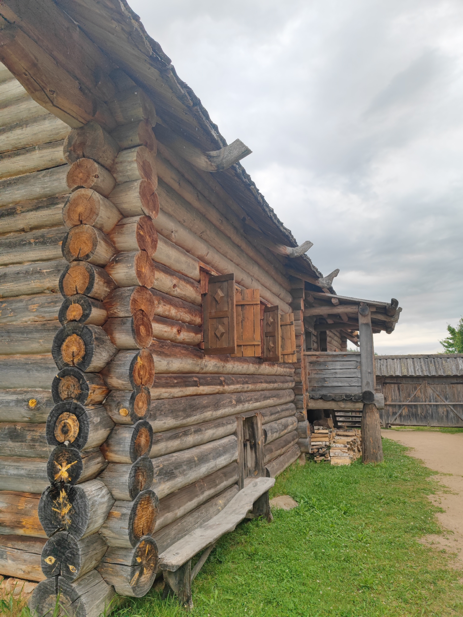 A close-up, low-angle shot shows the side of a rustic log cabin in the village of Bugrovo, Russia. The cabin is constructed from weathered, horizontal logs, with a distinctive corner joint featuring vertical logs. Wooden shutters are closed on a small window. A rough-hewn wooden bench sits in front of the cabin, partially obscured by green grass. To the right, a wooden porch with a roof overhang supported by posts is visible, along with stacked firewood. Beyond the porch, a rustic wooden fence and gate are present, bordering a dirt path. The sky above is overcast with grey clouds.
