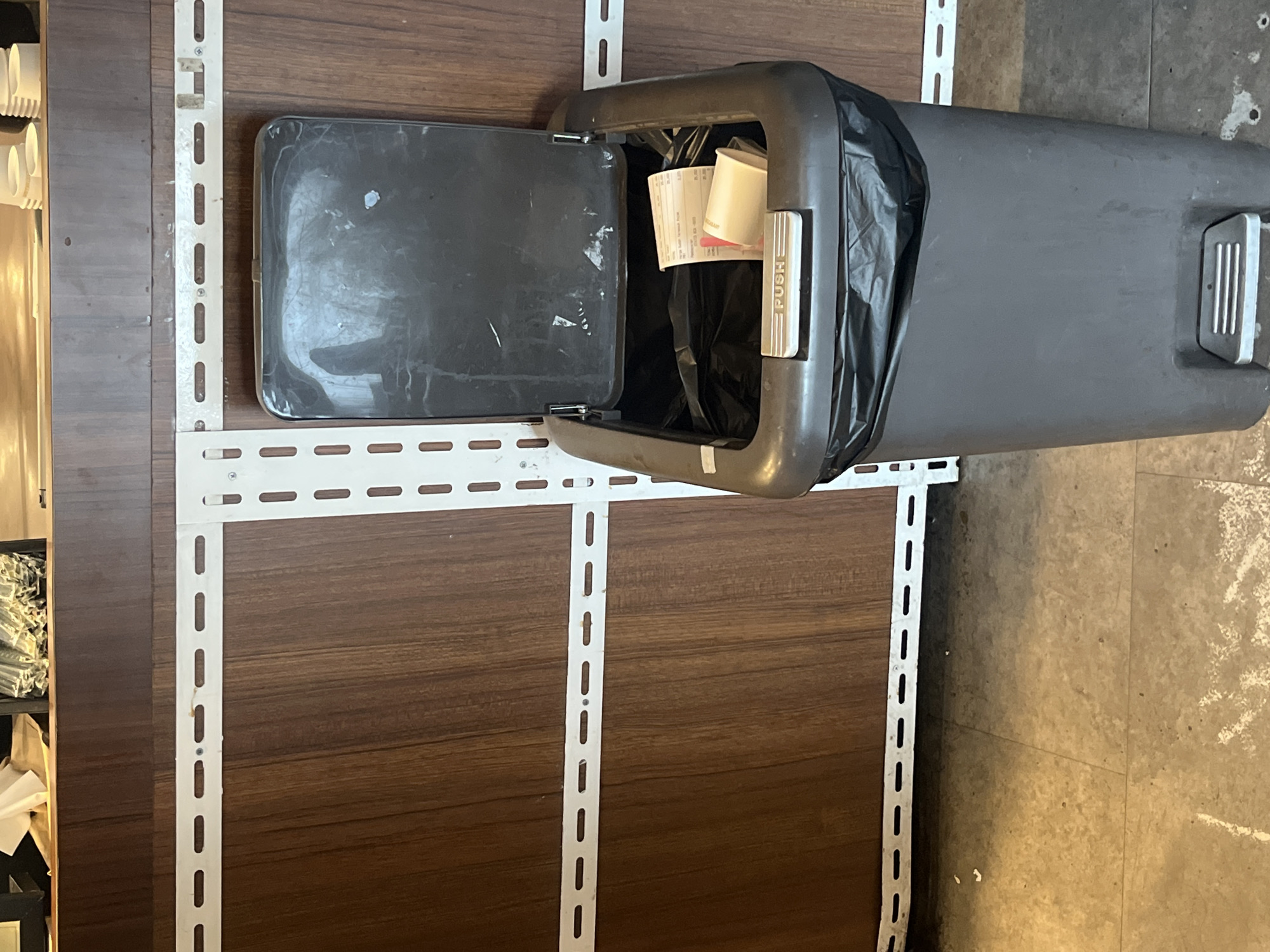 The image shows a dark grey pedal trash can with its lid open, positioned against a wooden wall. The trash can is lined with a black plastic bag, and inside, there are some receipts and a small white paper cup. A metallic "PUSH" pedal is visible on the front of the trash can. The wall behind the trash can has a wood-grain pattern and is decorated with white horizontal metal strips with oblong holes. The floor in front of the trash can is tiled in a neutral color, with some white marks on it. There are no people or animals visible in the image. The scene appears to be indoors, possibly in a commercial establishment. The provided context of West Jakarta, Indonesia is not visually verifiable from the image.