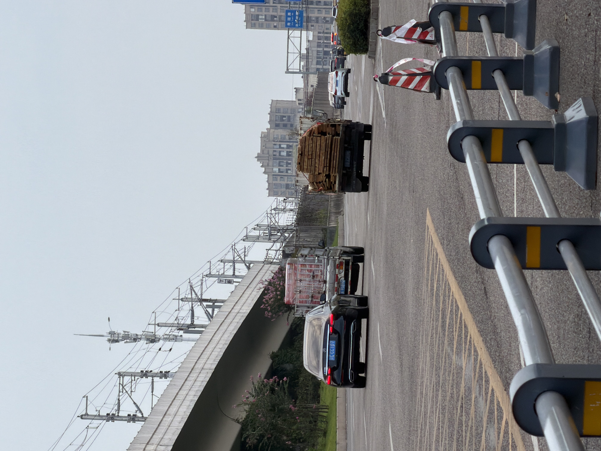 The image depicts a street scene in Jiangjin, China. In the foreground, a gray metal barrier with yellow markings separates the viewer from the road. Traffic cones with red and white stripes are placed on the right side of the road. In the middle ground, a black Mercedes-Benz sedan is driving on the road, followed by a flatbed truck carrying lumber. Further down the road, several other vehicles are visible, including a white Audi and a silver SUV. In the background, tall buildings and power lines are visible against a cloudy sky. The overall impression is a typical urban street scene with ongoing traffic and infrastructure.