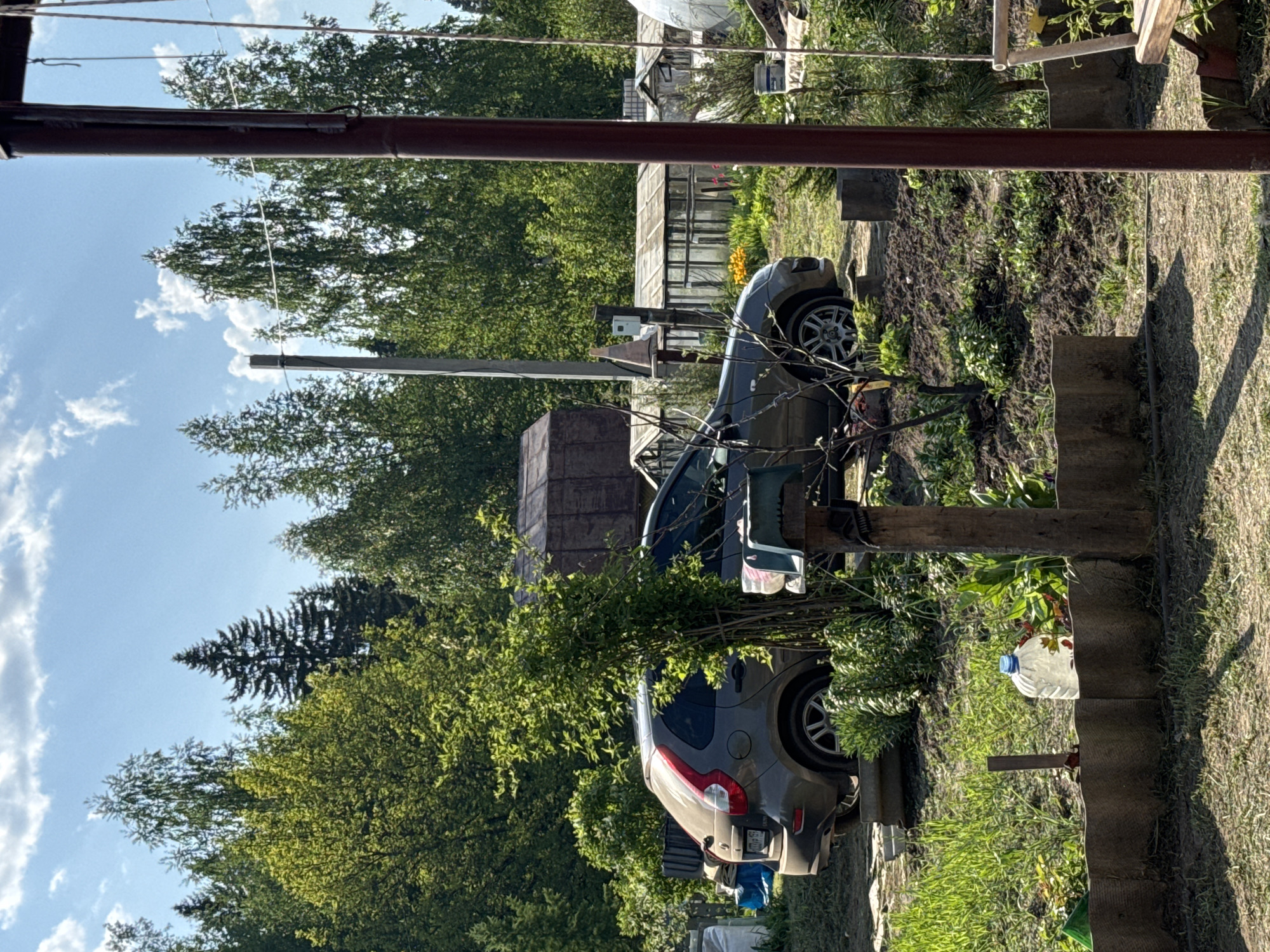 The image depicts a gray Volvo XC60 parked in a yard in what appears to be a rural or semi-rural setting. The car is surrounded by greenery, including trees, bushes, and grass. In the background, there are structures that could be houses, sheds, or greenhouses. The sky is visible with some clouds. A few objects appear to be related to gardening. The photograph appears to be taken during daylight hours. There is no evidence of any people in the immediate view. The location is Krasnokamsk, Russia, though that is not evident from the image itself.