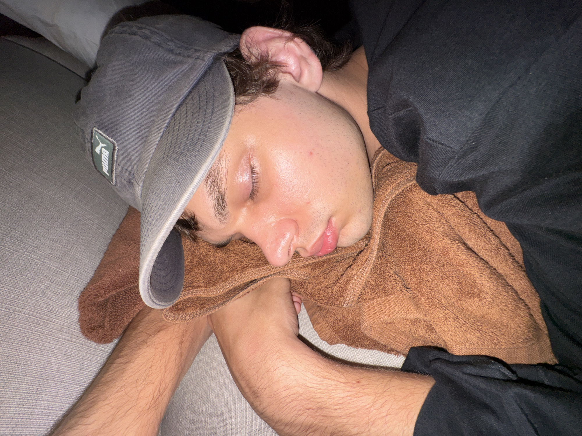 A person is asleep on a gray upholstered surface. They are wearing a gray Puma baseball cap, a black hooded sweatshirt, and resting their head on a brown towel. Their arms are crossed underneath the towel. The scene appears to be indoors. The location in Kuta Selatan, Indonesia cannot be determined from this image alone.