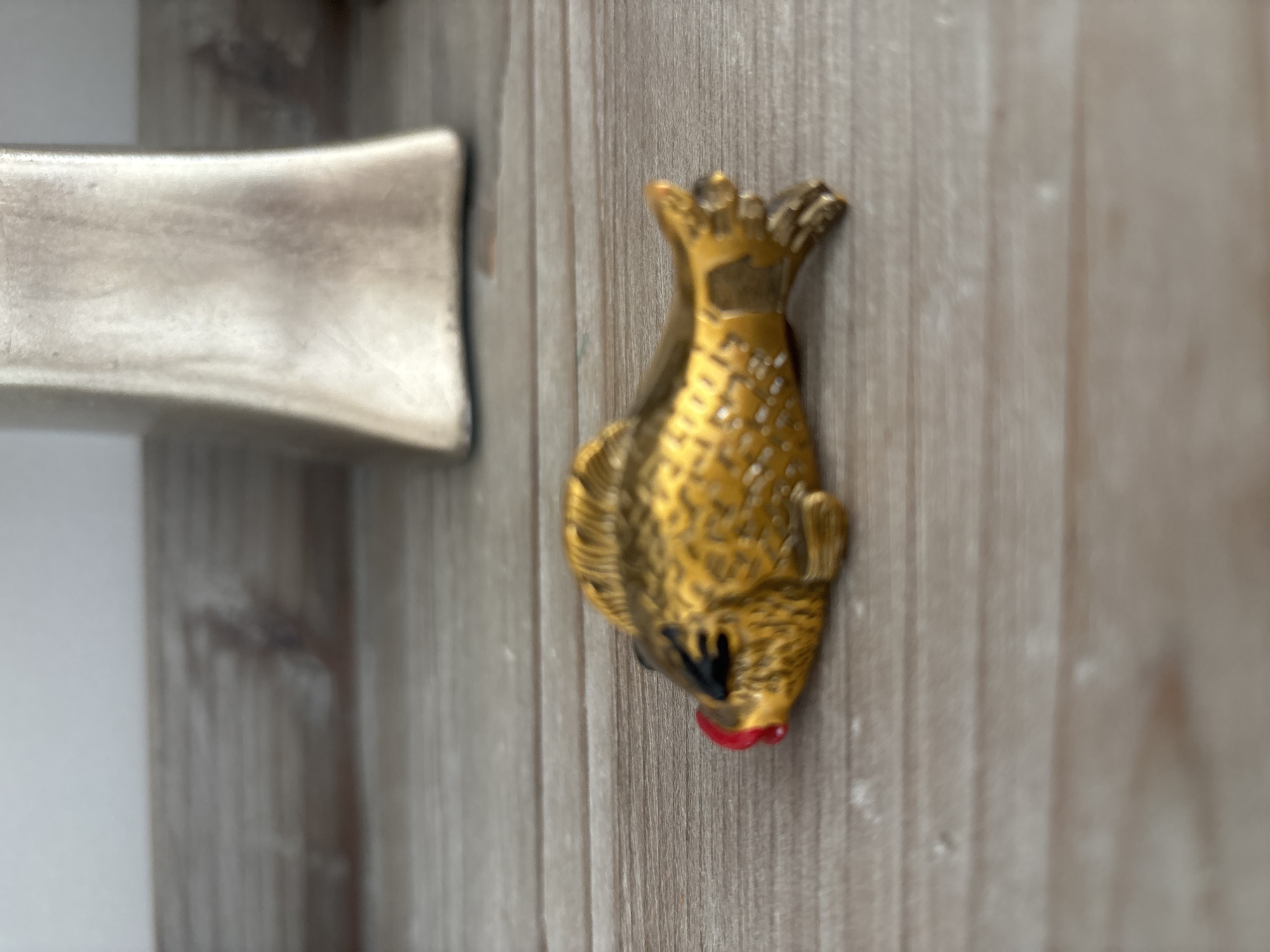 The image shows a close-up, side view of a small, golden, decorative fish ornament lying on a wooden surface. The fish has black eyes and bright red lips. In the blurred background, a tall, metallic, rectangular object, possibly a vase or sculpture, is visible. The wooden surface appears to be a shelf or tabletop. There is no indication of the location in Dubai, United Arab Emirates.