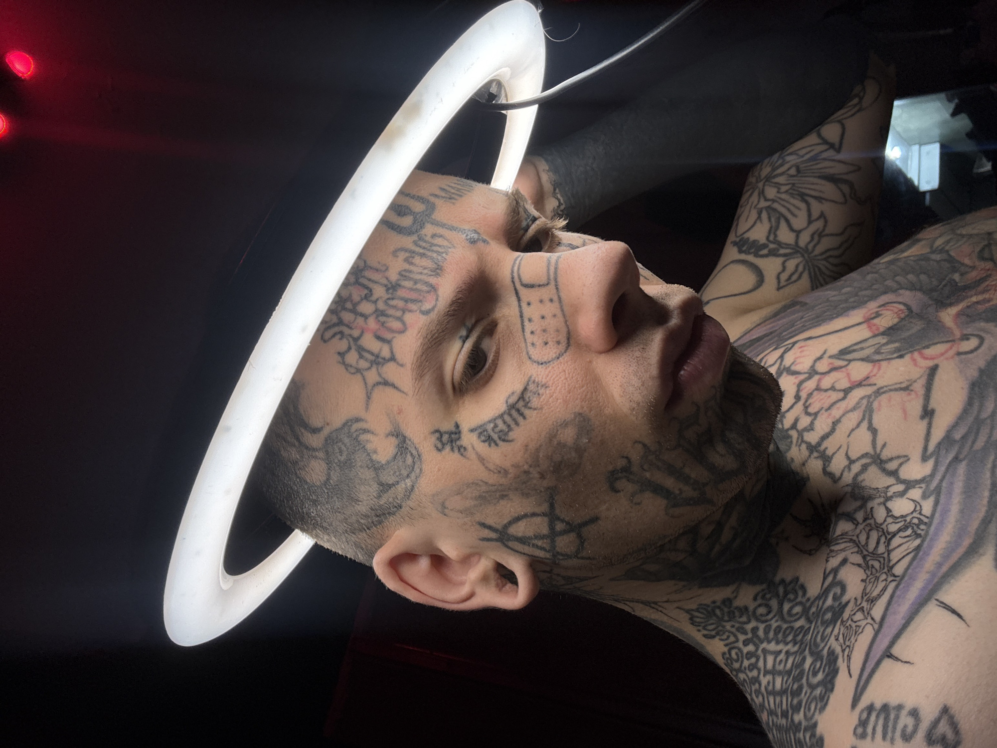 A heavily tattooed young adult male is captured in a close-up portrait, likely in a dimly lit indoor setting, possibly a studio. He has light skin, light-colored eyes, and short hair or a shaved head with stubble on his chin and cheeks. His face, neck, and upper torso are extensively covered in various black ink tattoos.
He is positioned with his head tilted slightly to his right, looking upwards and to his right, with a neutral or contemplative expression. Directly above his head, a bright, circular white ring light casts a prominent glow, creating a halo effect and illuminating his face. A black cable is visible, connected to the ring light. The background is predominantly dark, almost black, with a subtle red glow originating from the top-left corner, hinting at another light source or indicator.
Notable details include the intricate and numerous tattoos. On his face, various script tattoos and symbols are visible, including a distinct outline of a bandage on his right cheekbone. Below his right eye, a tattoo in Devanagari script reads "अहं ब्रह्मास्मि" (Aham Brahmasmi), which translates to "I am Brahman." Other symbols, such as a trident-like design on his forehead and a cross-like symbol on his left cheek, are also present. His neck and arms display complex patterns, including floral designs on his left shoulder/arm and abstract or script tattoos, with partially visible text resembling "CLUB" on his lower right arm/shoulder area. The extensive tattooing is a central feature of the image. The artificial lighting suggests the photo was taken indoors, likely at night or in a controlled environment. The location context provided is Buenos Aires, Argentina, although no specific visual cues in the image confirm this.