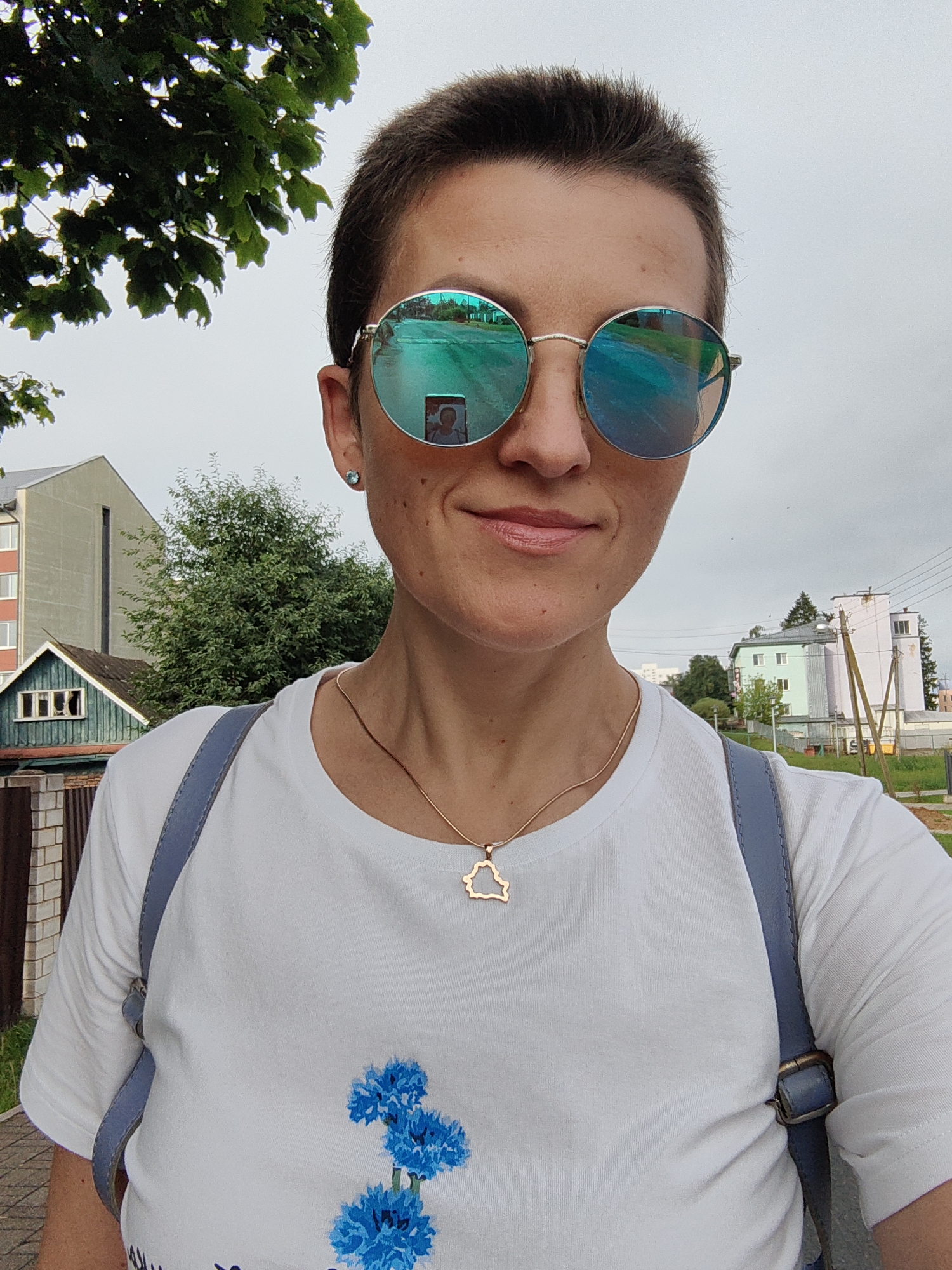 A woman with short brown hair and sunglasses is taking a selfie outdoors. She is wearing a white t-shirt with a blue floral design and a gold necklace with a pendant shaped like the outline of Belarus. Her backpack straps are visible over her shoulders. In the background, there are buildings, trees, and a cloudy sky, characteristic of the city of Fanipal, Belarus. The reflection in her sunglasses shows the surrounding environment, including a street and buildings.