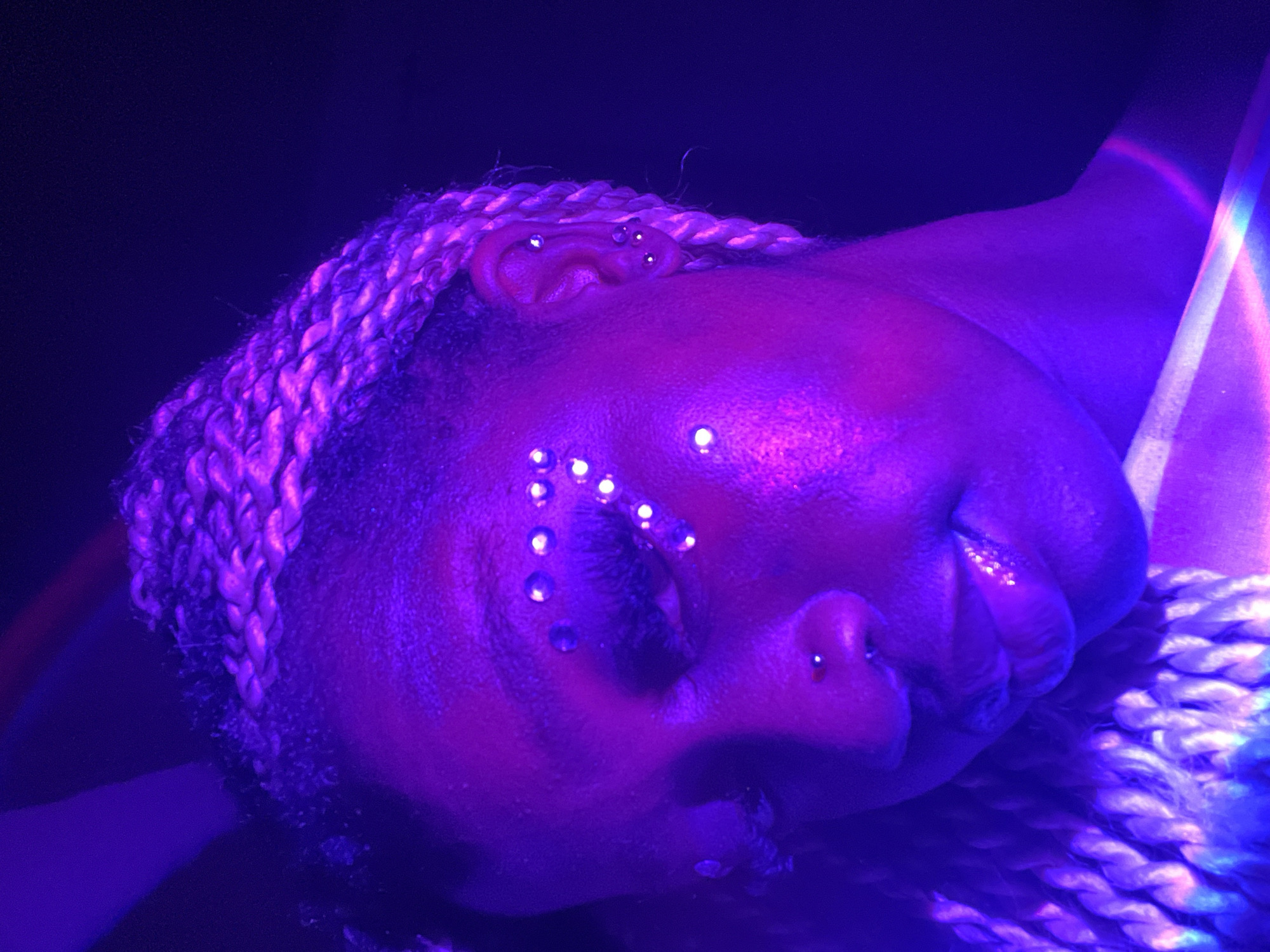 A close-up, tilted shot under predominantly purple lighting features a person's face. The individual has a dark complexion and their eyes are closed. A series of small, shiny dots are arranged around their left eye. They have multiple piercings in their ear, and a piercing on the left side of their nose. Their hair is styled in blonde braids. The background is dark and indistinct. A bright, multi-colored light streak is visible on the right side of the image. The image does not contain enough information to determine the exact location within Ado, Nigeria, or specific actions, interactions, or additional objects within the environment.