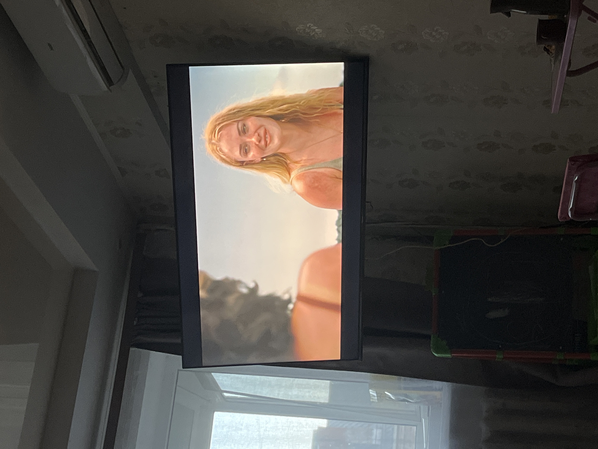 The image depicts a television screen displaying a scene of a blonde woman smiling. The setting appears to be outdoors, possibly a beach. The TV is mounted on a wall in a room, positioned above a window. To the left of the TV is a white air conditioning unit. A child's easel and chair are partially visible on the right side of the frame. The wallpaper is patterned. There is no information to verify that the location is Izhevsk, Russia.