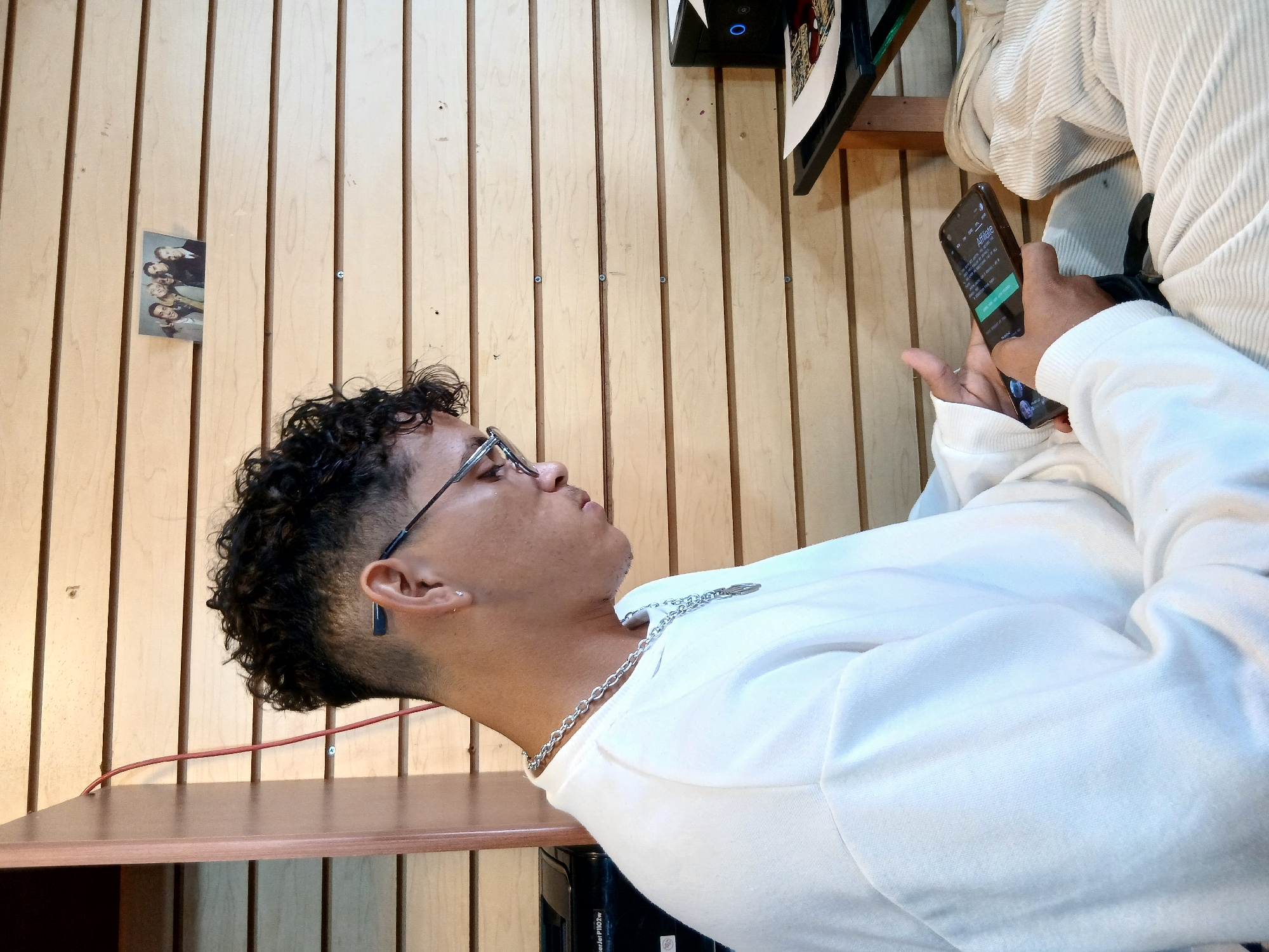 The image, oriented correctly, shows a young man intently looking at a smartphone held in his right hand. He appears to be in his late teens or twenties, with curly dark hair, wearing clear-framed glasses, a white long-sleeved shirt, and a silver chain necklace. A dark, flat rectangular earring is visible in his left ear. His expression is focused.
The background consists of a light-colored wooden slatted wall. A dark wooden shelf or table extends across the lower portion of the frame, with a red electrical cable running along the wall above it. On the wooden wall, a small, partially visible picture of a group of people is pinned. Higher up, a dark, framed object, possibly a monitor or artwork, is also visible. White, ribbed fabric, which could be part of upholstery or another garment, is present in the upper right corner of the background. Several screws are visible securing the wooden slats.
On the illuminated screen of the smartphone, the word "Afilia" is visible at the top, along with other text and a prominent green button, indicating an app or website interface. The setting is an indoor space, suggestive of a casual environment, located in Juan Griego, Venezuela.