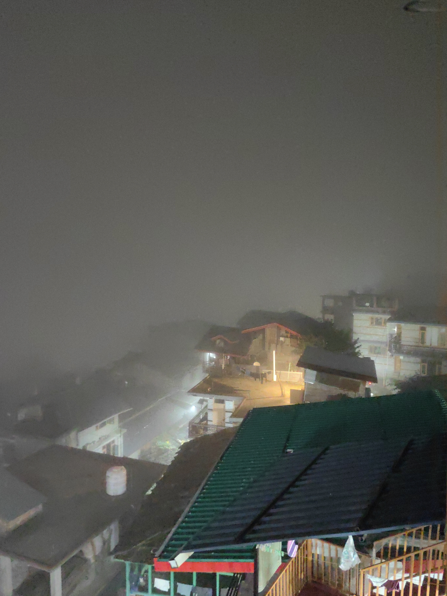 The image depicts a nighttime view of a hillside village, likely Vashisht, India, shrouded in heavy fog. The scene is illuminated by artificial lights, casting a dim glow on the structures. Multiple buildings with sloped roofs are visible, some constructed with light-colored walls and dark trim, others with darker roofing materials. A prominent green, corrugated metal roof dominates the foreground, with a wooden railing and stairs leading to a lower level. A white water tank sits on another rooftop to the left. The fog obscures distant views and creates a diffused atmosphere. There are no people clearly visible in the image.