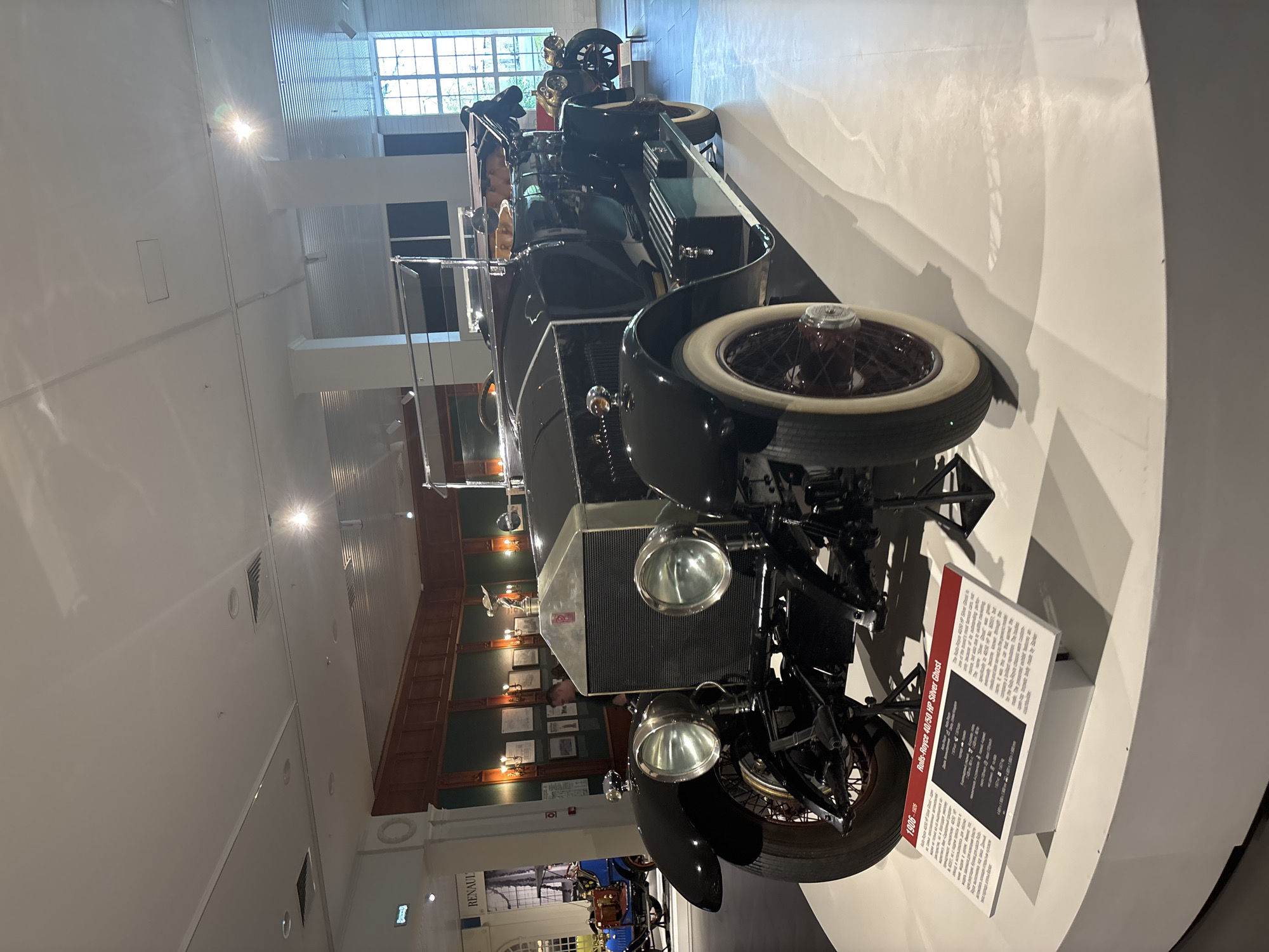 The image shows a black 1908 Rolls-Royce 40/50 HP Silver Ghost on display in a museum. The car is positioned on a white circular platform. In the background, there are other vintage vehicles, including a blue car, and displays with historical information. The museum has wooden paneling on the walls and large windows in the background. The scene appears to be well-lit with overhead lighting. The car is the primary subject, and its details, such as the large headlights, prominent grille, and spoked wheels, are clearly visible. There is a descriptive plaque in front of the car, written in Russian and English, identifying the vehicle and providing specifications. A person is partially visible in the background, standing near another display. The location is the UMMC Museum of Military and Automotive Technology in Verkhnyaya Pyshma, Russia, as indicated by the context of the collection.