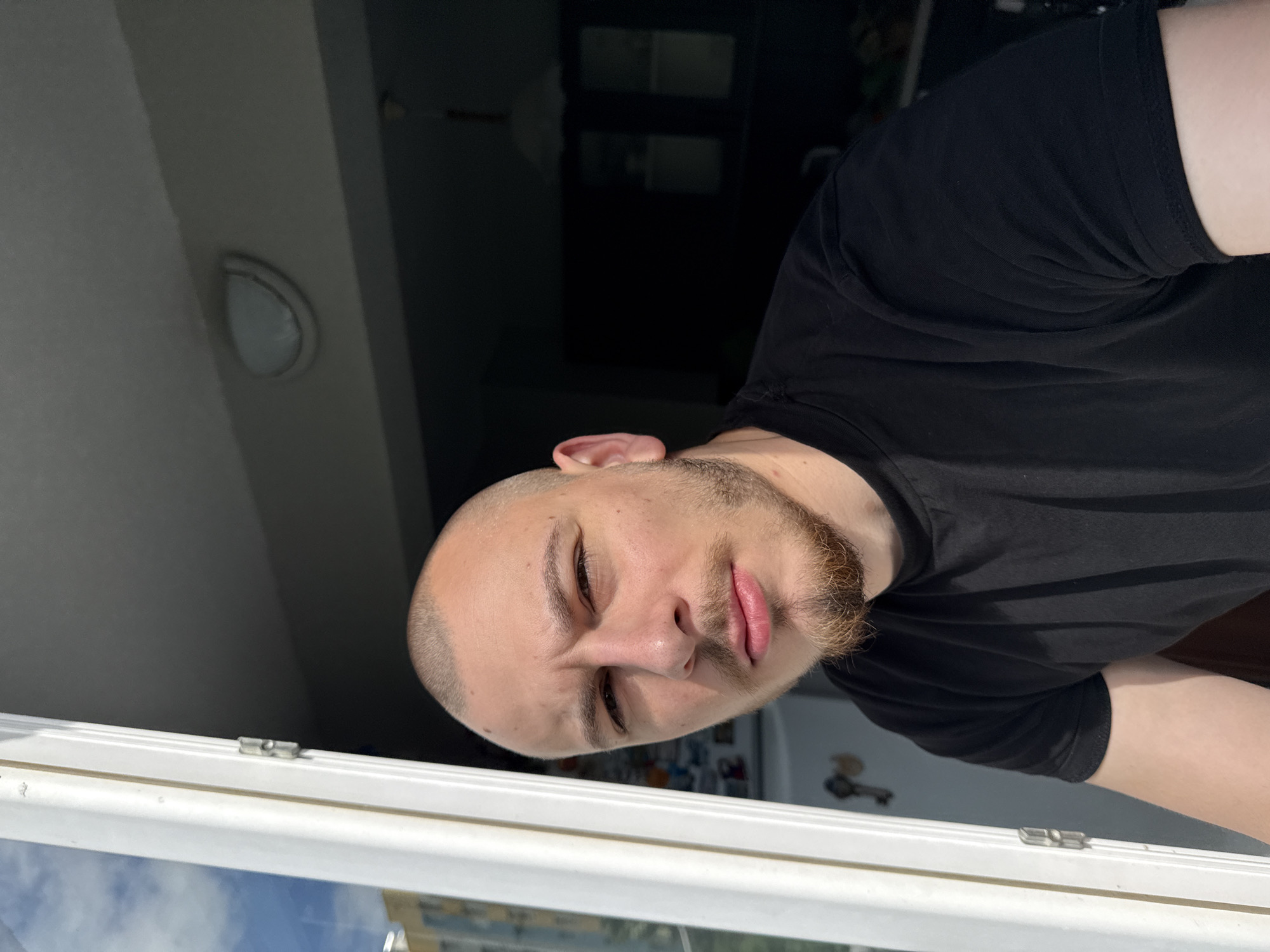 A bald man with a beard is looking at the camera. The man is wearing a black shirt. He is standing in front of a window, through which a building is visible. The location is Stara Zagora, Bulgaria. There is a ceiling light in the background.