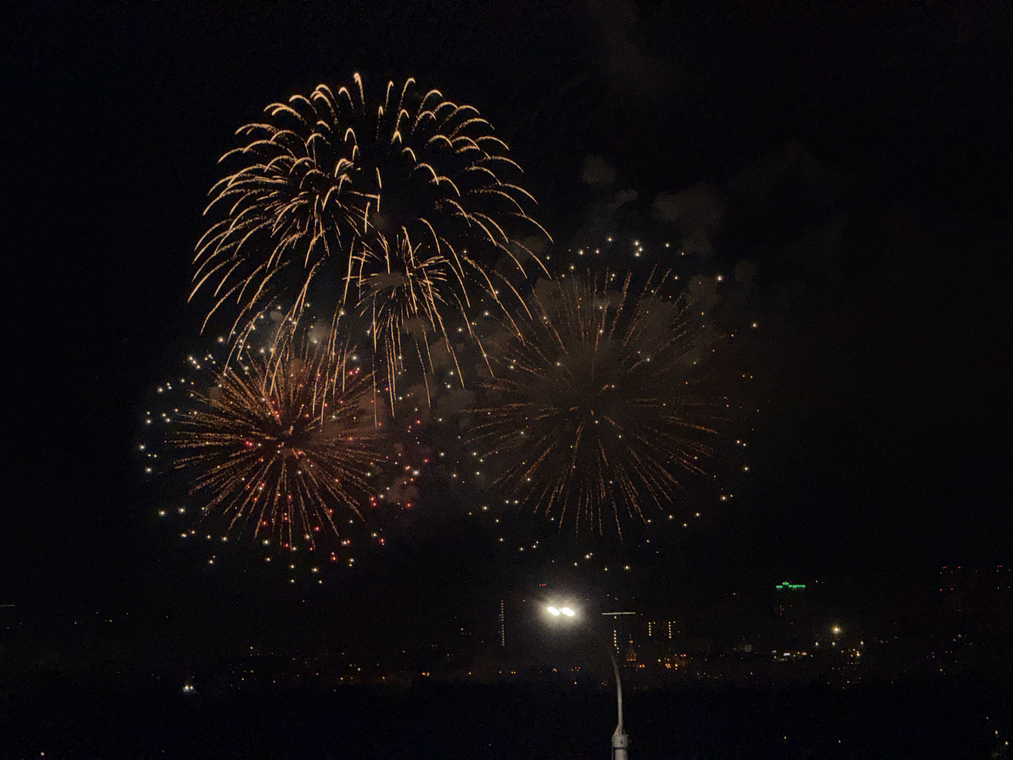The image depicts a nighttime scene with fireworks in the sky above a cityscape. Three large bursts of fireworks are prominent, with varying patterns and colors ranging from gold to red. The fireworks are set against a dark sky. Below the fireworks, a street lamp illuminates a portion of the scene, and in the distance, a city skyline is visible. The cityscape includes buildings of varying heights and lights. The location is likely Krasnoyarsk, Russia, based on the description prompt.