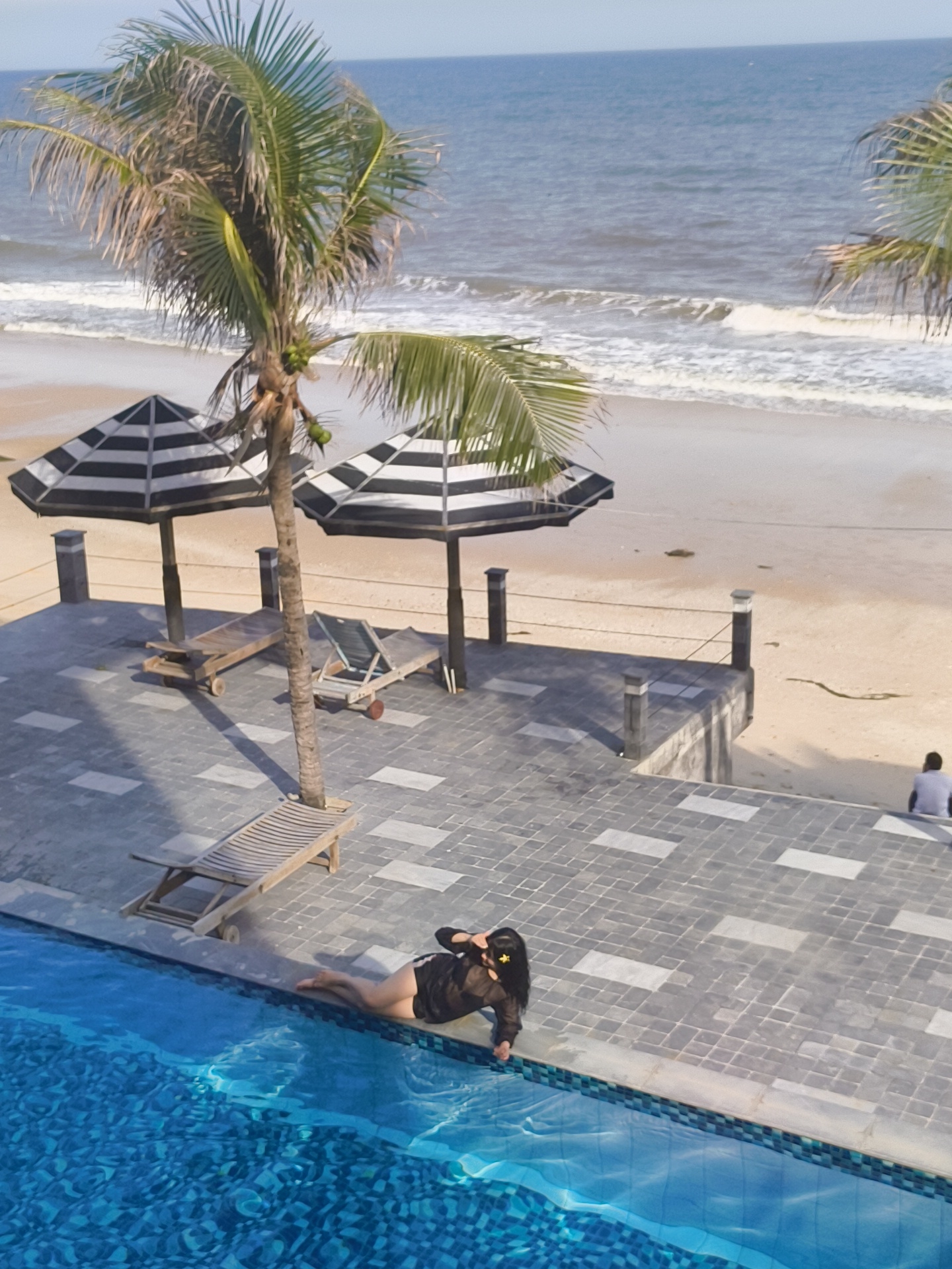 A woman is lying on the edge of a swimming pool overlooking a beach. The woman, wearing a black translucent cover-up and dark shorts, is on her left side with her head resting on her left arm. She has dark hair and is wearing yellow flower accessories in her hair. The swimming pool is in the foreground with blue water. Behind the pool is a tiled patio with several wooden lounge chairs and two striped umbrellas. A palm tree stands near the lounge chairs. Beyond the patio is a sandy beach with gentle waves breaking on the shore. In the distance, a person is sitting on steps facing the beach and ocean. The sky is clear and blue. This scene appears to be a resort or hotel in Phu Thuy Ward, Vietnam, given the tropical vegetation and beach setting.