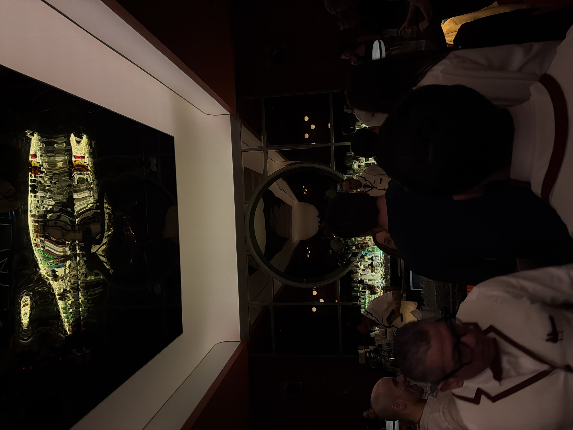 This photograph captures the interior of a bar, likely located in Mexico City, Mexico. A group of people are gathered at the bar, with some facing the bartender, visible in the background, and others facing the camera. The scene is dimly lit, with the interior decor featuring dark tones and the warm glow of the bar lighting. A prominent architectural feature includes a circular window or opening in the back of the bar, revealing the city lights. A glossy, dark surface reflects the bottles and lights from the bar, creating a distorted reflection. The individuals appear to be engaged in conversation and interaction, suggesting a social setting. The details of clothing are visible, showcasing casual attire with white and dark-colored shirts.