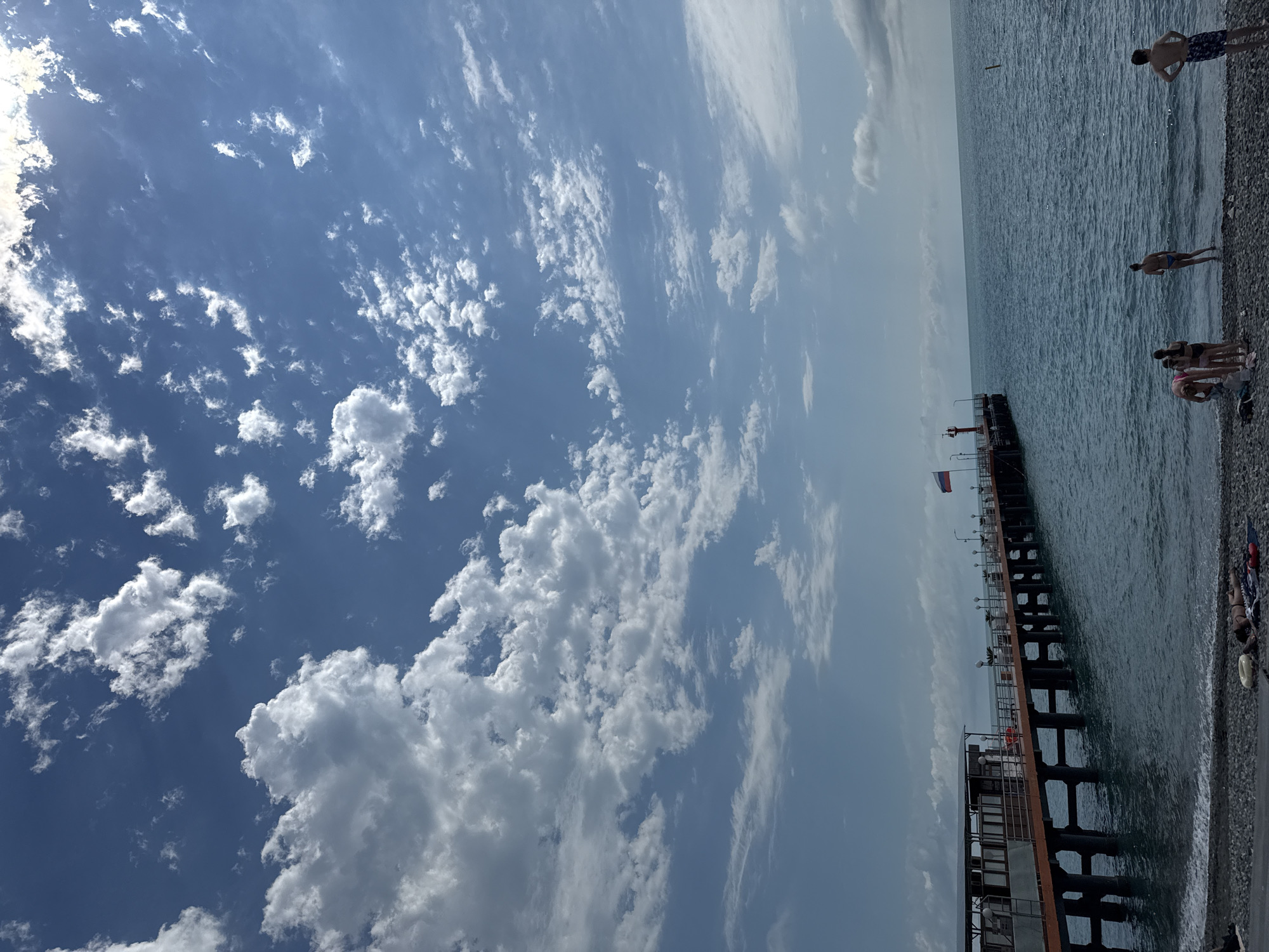 The image depicts a beach scene in Sochi, Russia, under a bright, cloudy sky. A long, dark pier extends into the rippling blue water. Several people are visible on the beach and near the water. On the far right of the image, one person stands on the shore, and further down, a few people are gathered near the water's edge. A person is wading in the shallow water.
The pier has a railing and lights along its length. A Russian flag flies from a pole on the pier. The beach is covered with dark pebbles. The water appears calm with small waves lapping at the shore. The sky is a vibrant blue with scattered white clouds, suggesting it is daytime. The overall atmosphere is relaxed, indicative of a day at the beach.