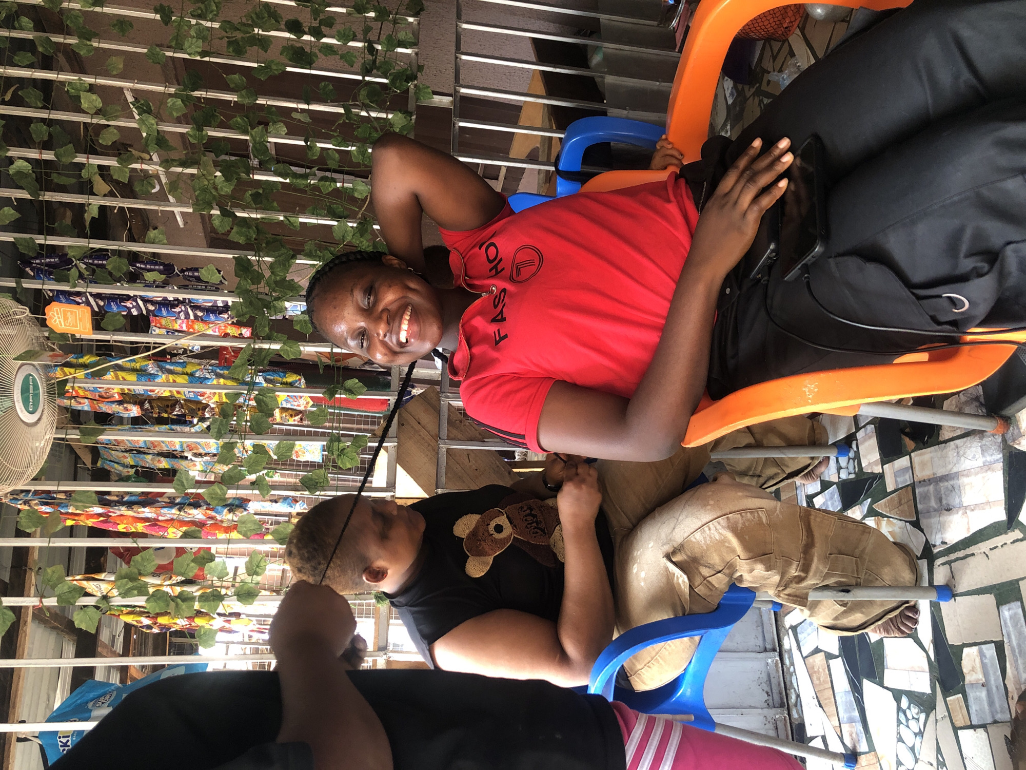 This image depicts a casual indoor or semi-outdoor setting, likely a small shop or waiting area, featuring three individuals.
The main subject is a young woman seated in an orange plastic chair on the left, smiling broadly and looking directly at the camera. She wears a red short-sleeved top with the word "FASHION" printed vertically on the front, along with a circular logo. Her hair is styled in braids. She has a black bag or fabric on her lap, with a black smartphone resting on it.
To her right, another individual is seated in a blue plastic chair, partially obscured. This person is wearing a black t-shirt with a brown teddy bear design on the chest and khaki-colored pants. They appear to be looking downwards or engaged with something in their lap. A third person is partially visible in the bottom left, wearing a black top and pink and white striped bottoms.
The background consists of white metal grates or shelving, draped with artificial green leafy vines. Behind these, shelves are stocked with numerous packaged goods, appearing to be snacks or drinks, forming colorful rows. A white electric fan is visible on the far left of the frame, above some products. An orange tag labeled "Surprise" hangs near the fan. The floor features a mosaic pattern made of irregularly shaped tiles in shades of grey, brown, and white. The overall impression is one of relaxation and a friendly atmosphere.