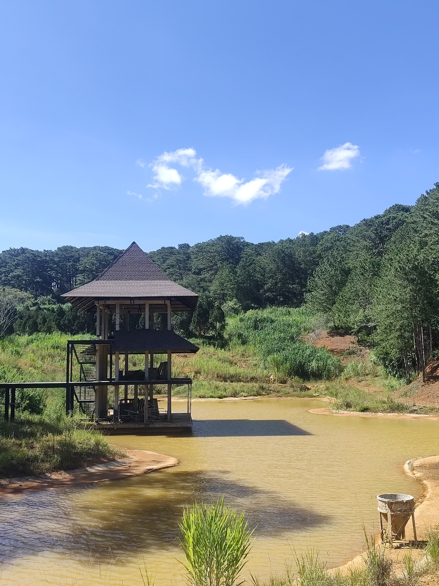 This image shows a wooden pavilion on a body of water, with a forested hill in the background. The pavilion has a dark, multi-tiered roof and is supported by white pillars. It appears to be on a wooden platform that extends into the water. The water is a murky, yellowish-brown color. The hill behind the pavilion is covered in dense, dark green pine trees. The sky is a clear blue with a few scattered white clouds. The overall scene suggests a natural setting, possibly a park or a recreational area in Da Lat, Vietnam, known for its pine forests and lakes. There are no people visible in the image.