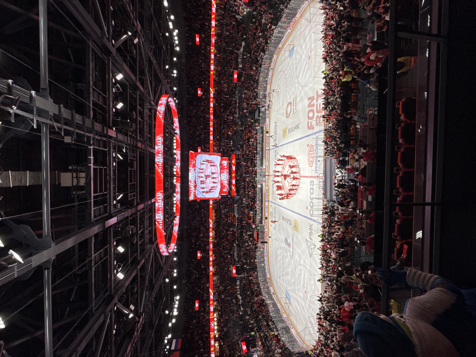 This image captures an overhead view of the Uralets Arena in Yekaterinburg, Russia, during an event. The scene depicts a brightly lit ice hockey rink surrounded by tiered seating packed with thousands of spectators, many wearing red and white attire and raising their hands in a gesture of enthusiasm. On the ice, approximately 20 individuals, uniformly dressed in red and white, form a compact circle near center ice, each with their arms raised overhead in a synchronized pose. The ice surface displays various sponsor logos including "LADA," "FONBET," "PMK," "CORAL," and "KHL" in Russian Cyrillic. Directly above the rink, a large rectangular jumbotron hangs, displaying an image of the group on the ice along with the Cyrillic text "ПОБЕДА!" (Victory!). This jumbotron is encircled by a dynamic circular LED display, predominantly showing red and white patterns. The arena's dark, industrial-style ceiling structure, complete with trusses and numerous lighting fixtures, is visible above. A Russian flag is mounted high on the arena's left side. In the immediate foreground, from a higher vantage point, the back of a young person wearing a blue knitted hat, a light-colored jacket, and blue mittens is partially visible, with their hands raised, mirroring the crowd's excitement.