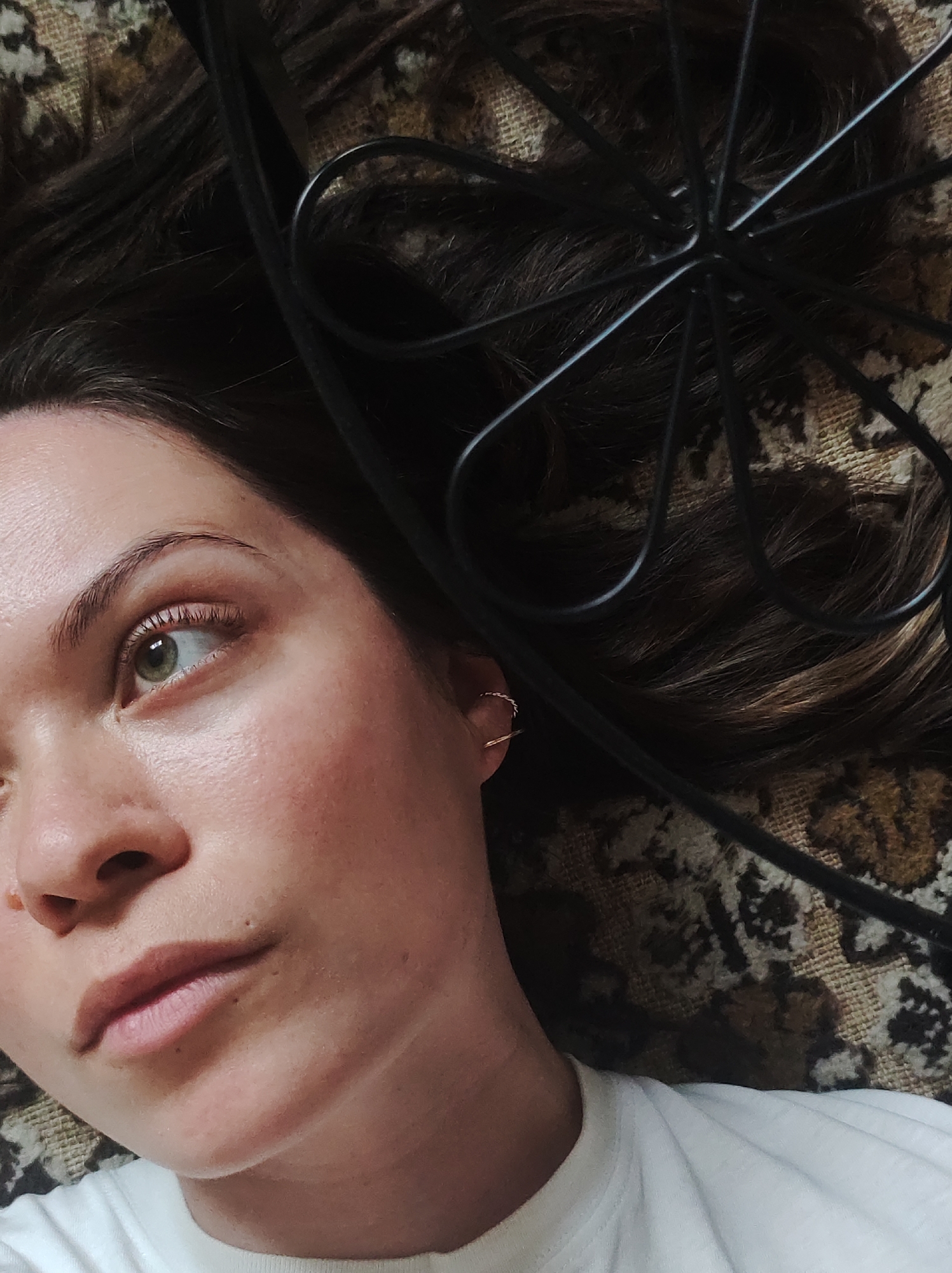 A woman with dark hair, light skin, and green eyes is lying on a patterned surface, possibly a rug or carpet. A black metal object, resembling part of a piece of furniture or a decorative item, is positioned near her head. She is wearing a white t-shirt and an earring in her left ear. The photograph is taken from above. The location is provided by the prompt as Фоки, Russia, although this information cannot be verified from the image itself.