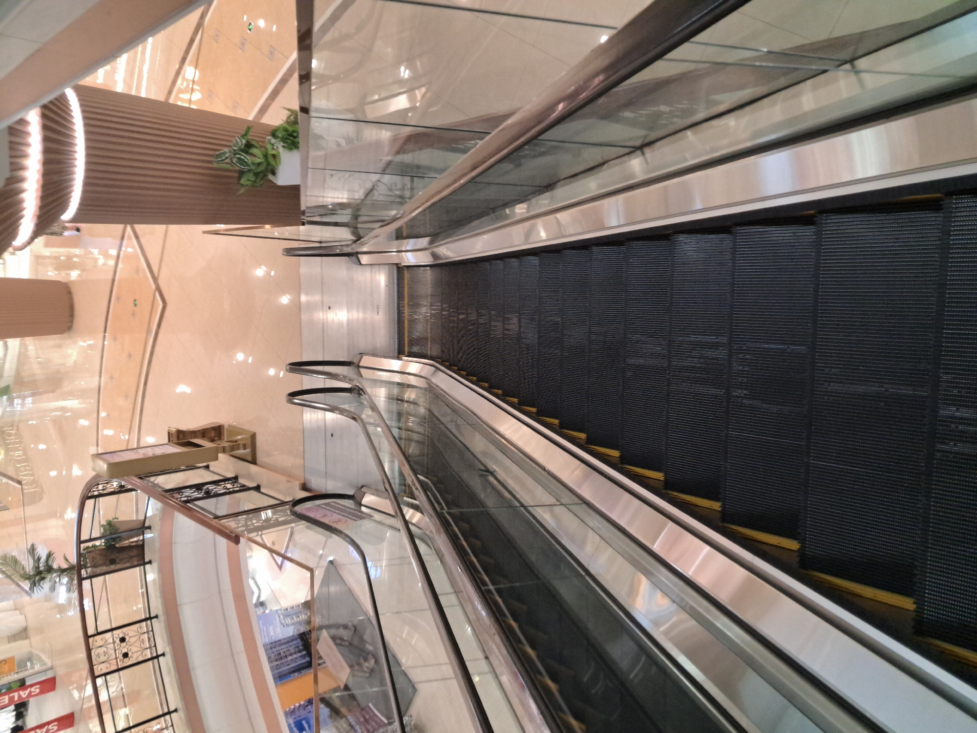 An overhead view of an escalator in a mall. The escalator steps are black with yellow edges and appear to be moving downwards. The handrails are metallic and the sides of the escalator are made of glass. In the background, the mall floor is visible, with a polished, reflective surface. There are decorative pillars and potted plants. No people are clearly visible on the escalator. The scene is well-lit, indicating it is daytime or the mall is brightly illuminated.