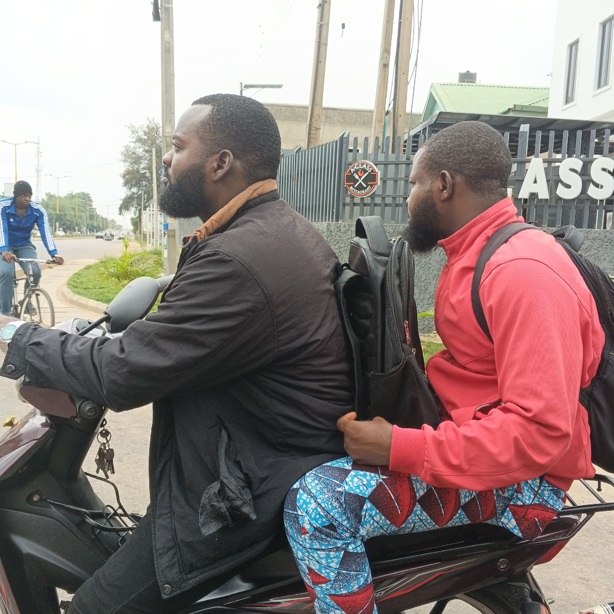 Two men are riding on a motorcycle, with the rider in the foreground and the passenger behind him. The rider is wearing a black jacket and black pants. The passenger is wearing a red jacket and patterned pants. Both men have backpacks. A third man is seen cycling in the background. The scene appears to be taking place on a street in Kabala, Nigeria, with buildings and trees visible. The weather seems overcast. Text visible on a sign in the background reads "A CLASS RESTAURANT".
