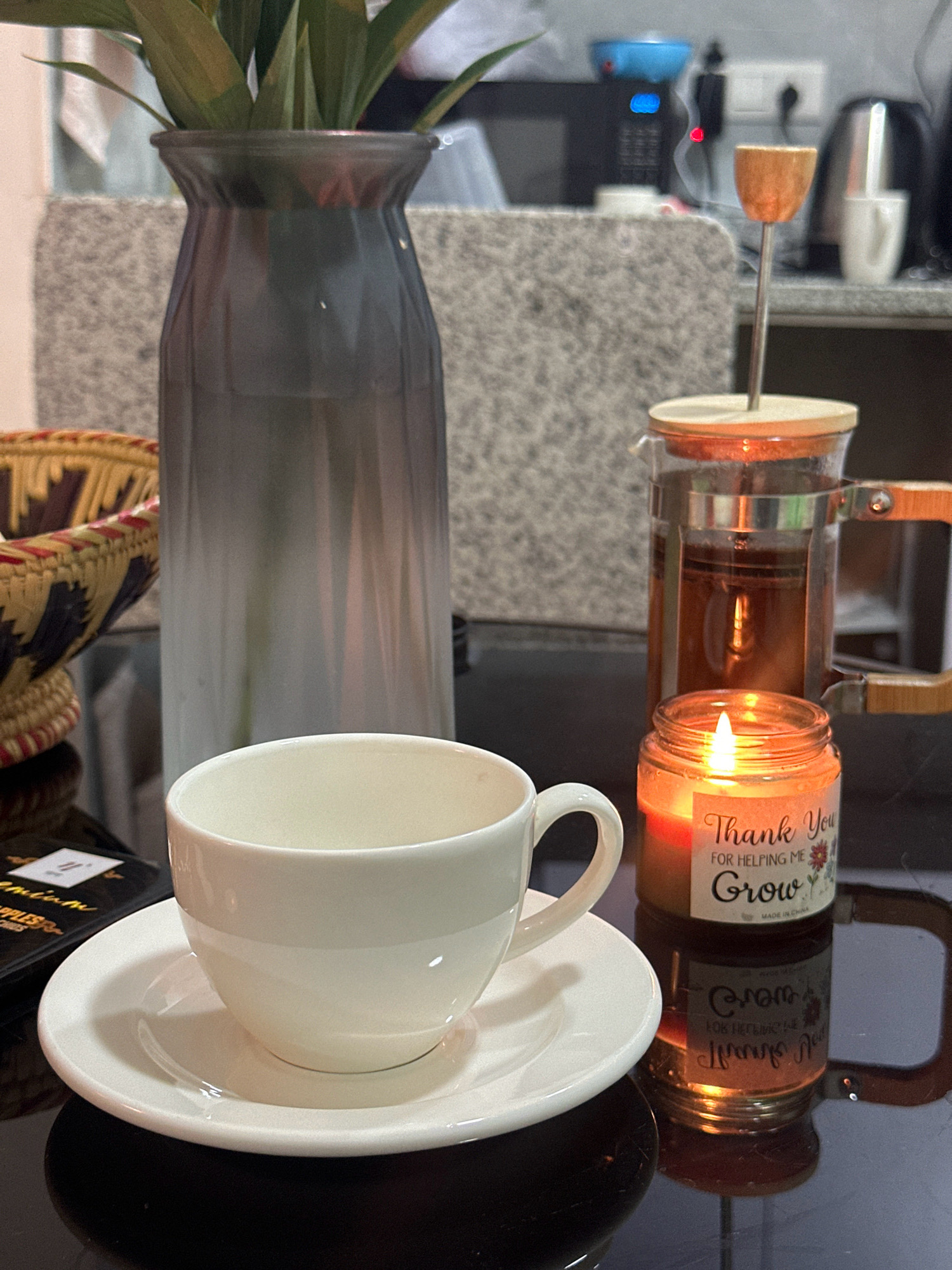 A close-up, eye-level shot shows a white teacup and saucer on a reflective black surface. To the right of the cup is a lit jar candle with the text "Thank You For Helping Me Grow" visible. Behind the candle and cup is a glass French press filled with tea, and further back is a tall, gradient-colored vase with green leaves. A woven basket is visible to the left of the vase. In the background, an electrical outlet and a kettle are discernible. The setting appears to be indoors, possibly in a kitchen or dining area, in Lalitpur, Nepal.
