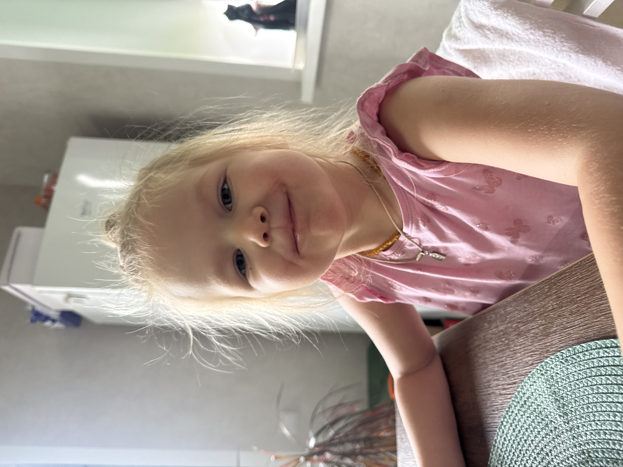 A young child with blonde hair, fair skin, and blue eyes is depicted in a close-up, slightly off-center portrait, smiling gently with a trace of a substance on the upper lip. The subject is seated at a wooden table within a domestic interior in the city of Ферма, Russia. The child wears a pink t-shirt with ruffled sleeves and glittery butterfly patterns, a light brown beaded necklace, and a silver pendant necklace. In the lower-left foreground, a teal green circular woven placemat is visible on the table. The background includes a window with a houseplant to the left of the child, and a white refrigerator with various objects on top to the right. Further to the right, another window features a dark, sculptural object on its sill, and a white towel is draped over a chair back next to the child. The scene is illuminated by natural light.