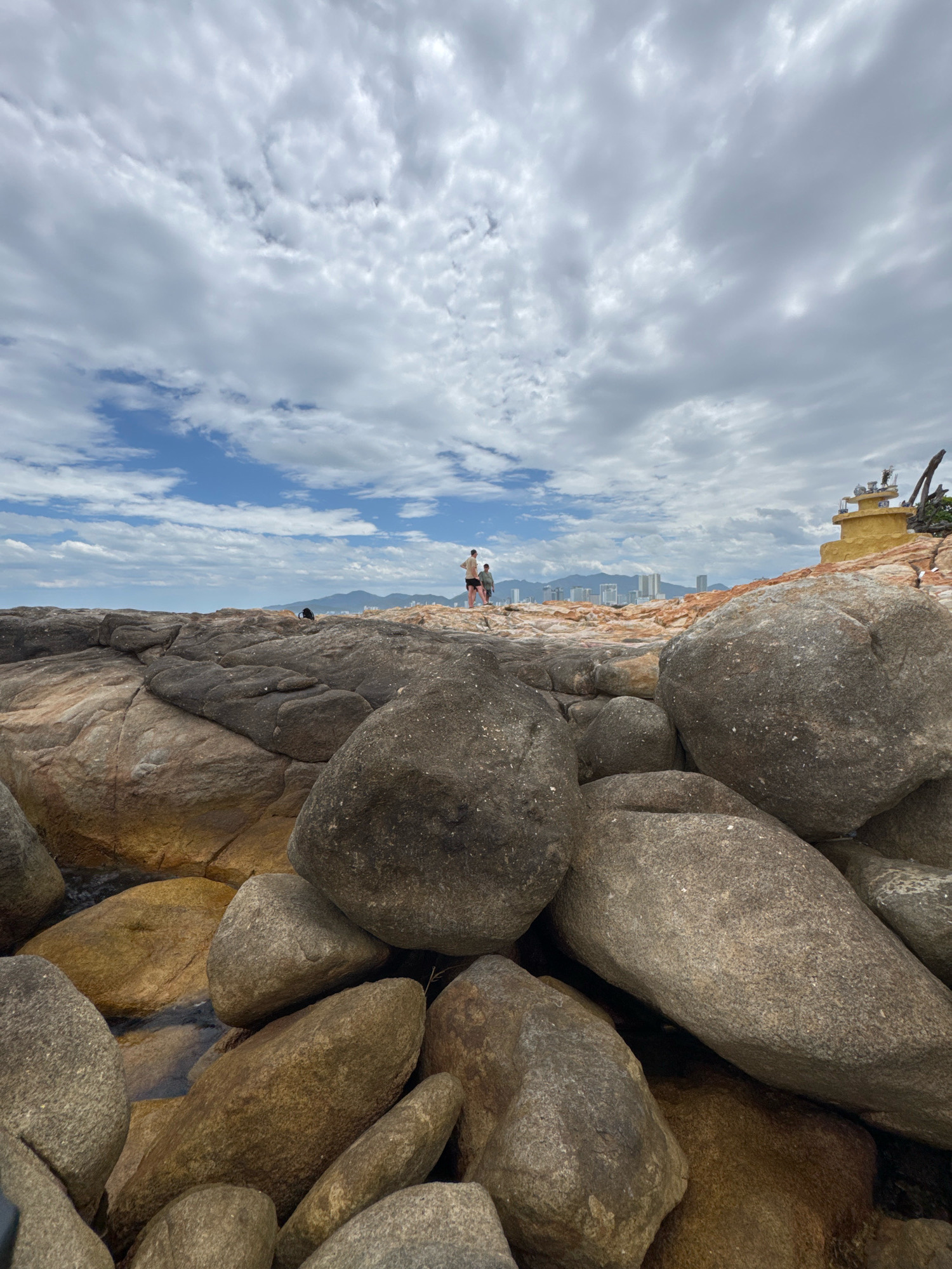 This is a photograph taken from a rocky coastline in Nha Trang, Vietnam. The foreground is dominated by large, rounded boulders of varying shades of gray and brown, some partially submerged in shallow, dark water. In the mid-ground, two people, a man and a child, stand on a rocky outcrop facing away from the camera. The man is wearing dark shorts and a light-colored shirt, and the child is wearing dark shorts and a light-colored top. Behind them, a cityscape with tall buildings is visible in the distance, nestled against a backdrop of rolling hills. The sky above is filled with a mixture of bright white clouds and patches of blue sky, suggesting a partly cloudy day. To the right of the frame, on a rocky incline, is a small, yellow, lighthouse-like structure with decorative elements on top, and a weathered piece of driftwood leaning against it. The overall scene depicts a natural coastal landscape meeting an urban environment under a dynamic sky.
