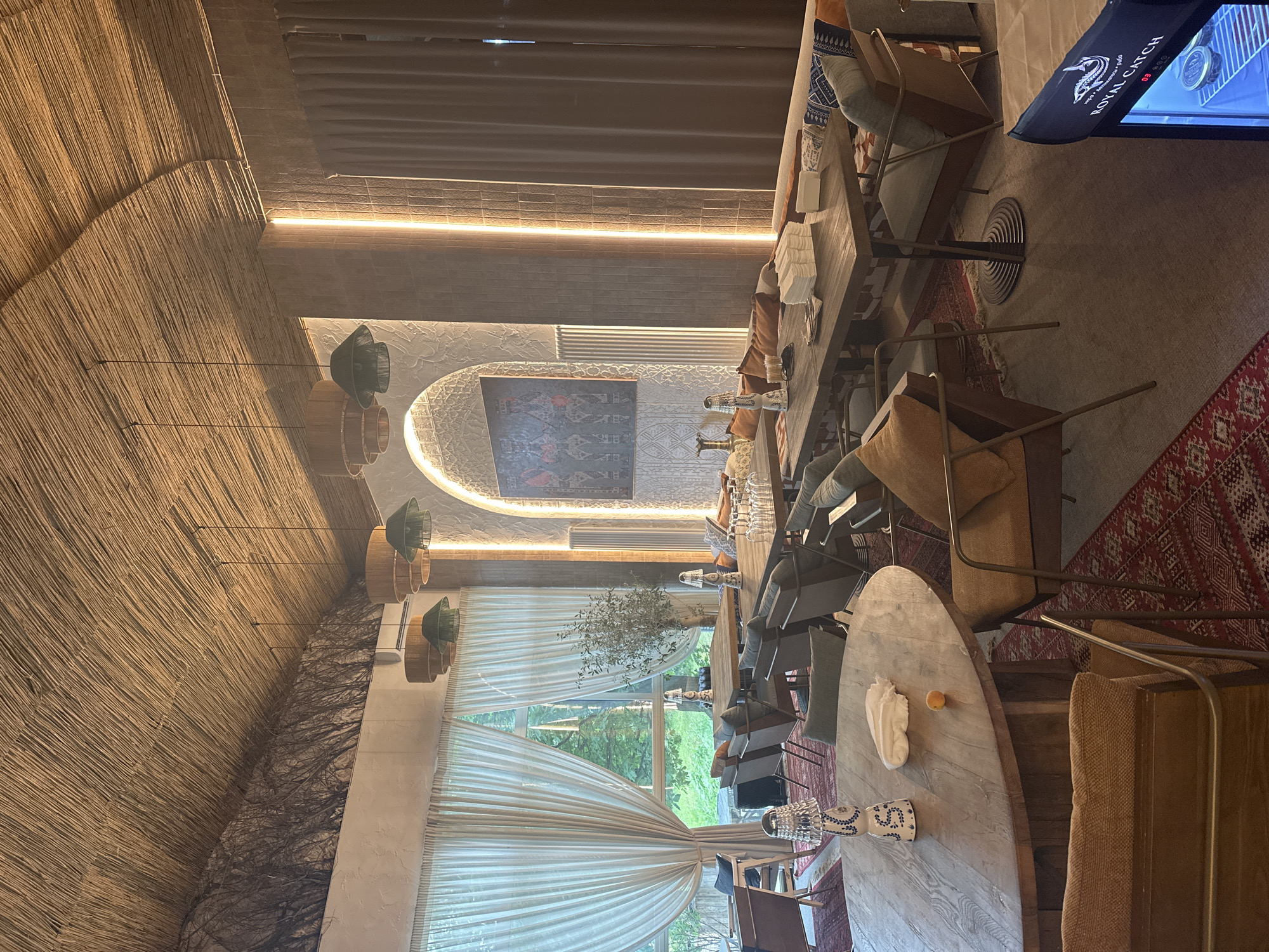 This is a medium shot of a restaurant interior in the city of Poselok Gidrouzla Imeni Kuybysheva, Russia. The restaurant features a rustic design with a thatched roof and natural wood furniture. Large windows with sheer curtains allow natural light to enter the space, and an assortment of pendant lights hang from the ceiling, casting a warm glow. There are several tables with chairs and lounge seating, arranged for dining. A large artwork hangs on the wall, and decorative elements such as lamps and plants add to the ambiance. The flooring is a mix of wood and patterned rugs. The scene is empty of people.