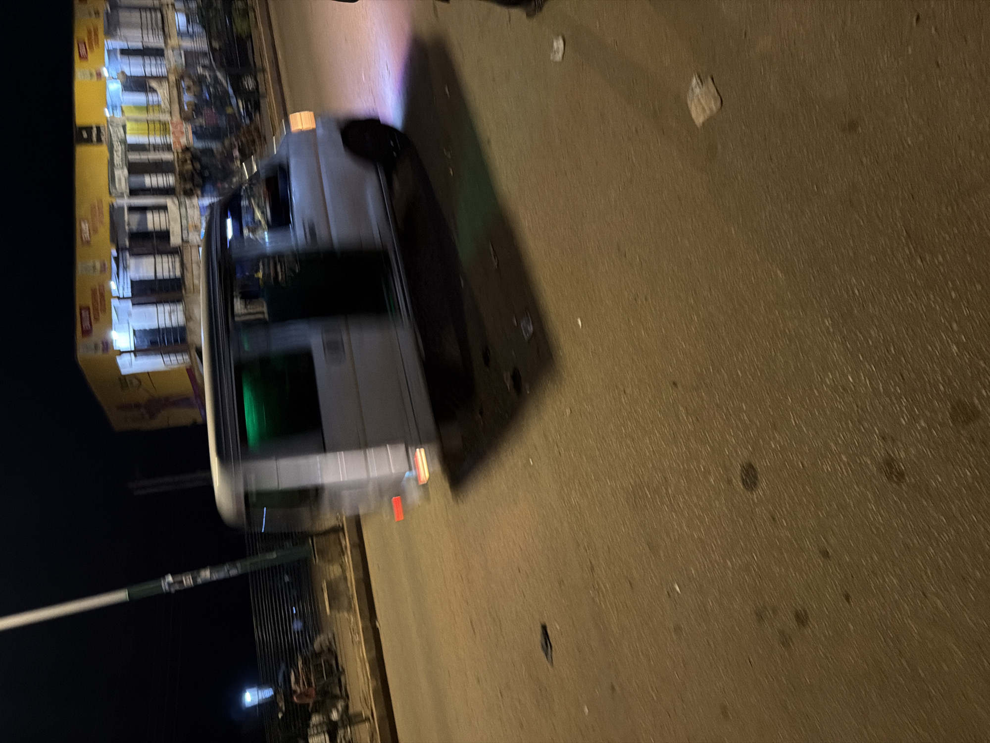 A silver van is captured in motion at night on a paved road in Bukuru, Nigeria, with its right side facing the viewer. The van, appearing to be a light commercial vehicle, exhibits motion blur. Its doors are open, and the windows are tinted green. The rear brake lights are illuminated red and orange, and a purple light casts a glow on the pavement from the right side of the vehicle. Trash and debris are scattered on the road surface. In the background, a two-story building with multiple windows and balconies is visible, illuminated by yellow and white lights from within and from signage above. A street light stands to the left of the frame, with advertisements attached to its pole. Several vehicles and a person pushing a cart are present on the sidewalk in front of the building. The scene is dark, with artificial lighting creating shadows.