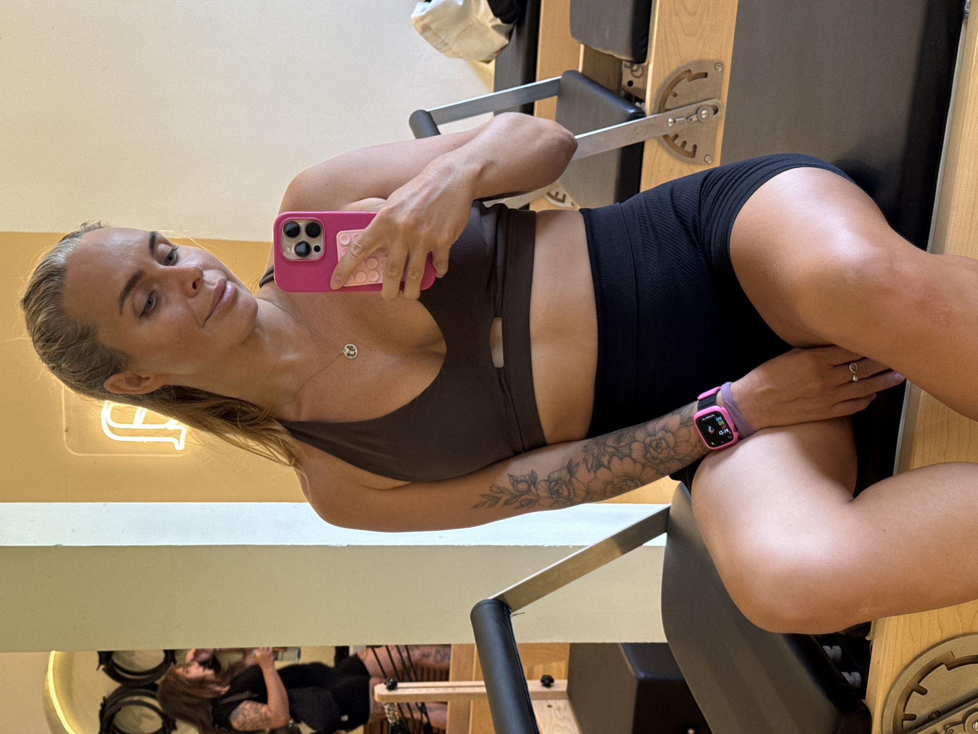 A blonde woman is taking a selfie in a gym. She is wearing a brown sports bra and black shorts. She has a tattoo on her arm and is wearing a pink smartwatch. In the background, another woman is visible, wearing a black top and shorts. The gym appears to have Pilates reformers. The scene is brightly lit.