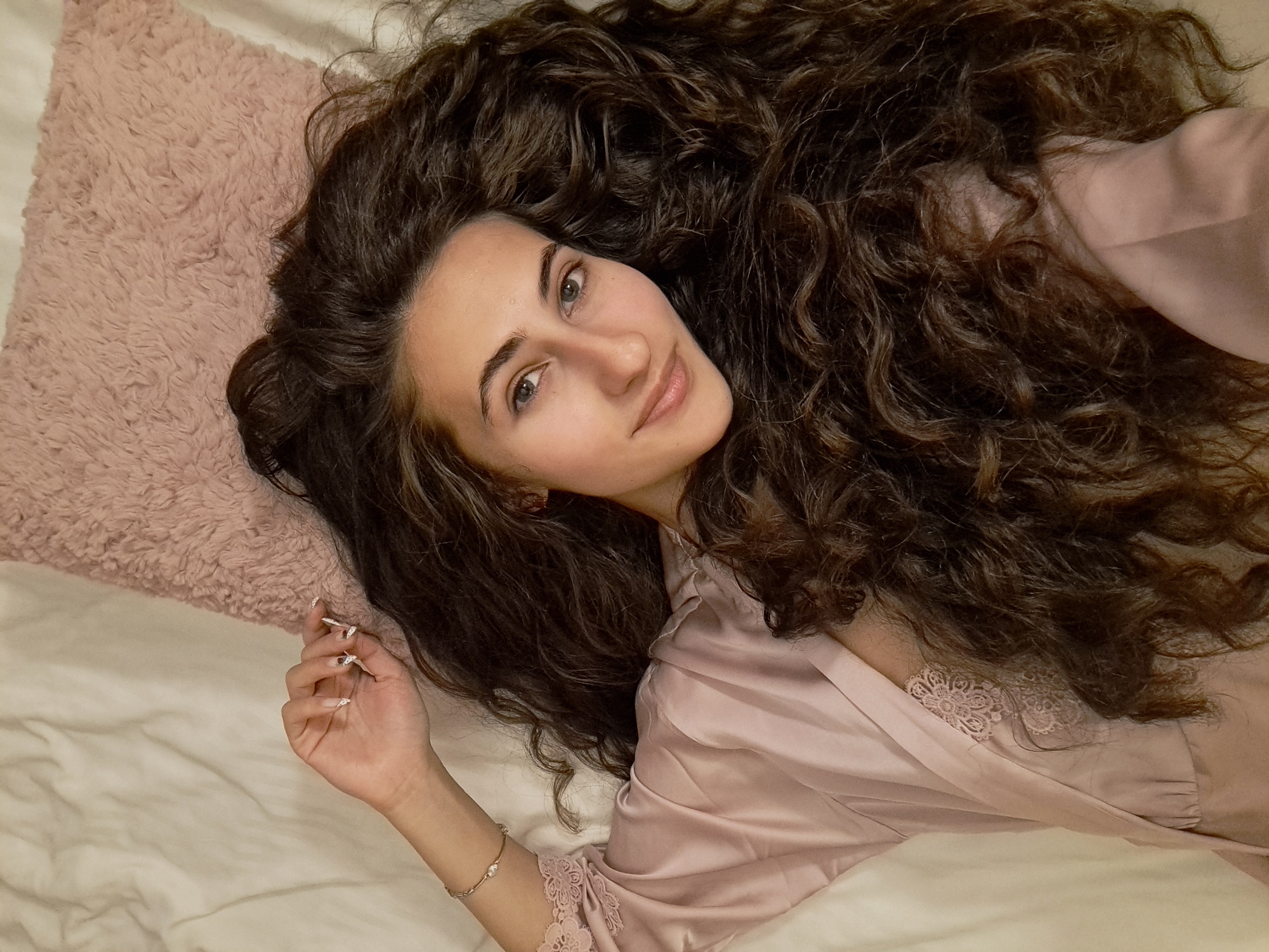 A young woman with long, voluminous, dark brown curly hair is pictured lying on a light-colored bed, her hair fanned out around her head. She looks directly at the camera with a calm, serene expression and a gentle smile. Her eyes are a light color. She is wearing a pale pink satin robe, which features delicate lace detailing on the sleeve and near the bodice. To her left, a soft, pale pink fluffy pillow is visible. A subtle, delicate bracelet adorns her left wrist, and her fingernails are adorned with a white and gold design. The setting appears to be an indoor space, likely a bedroom, characterized by the soft bedding. The lighting is soft, suggesting a relaxed atmosphere. There are no visible location cues for Negovan, Bulgaria, within the image itself, nor is there any visible text. The overall scene conveys a sense of comfort and tranquility.