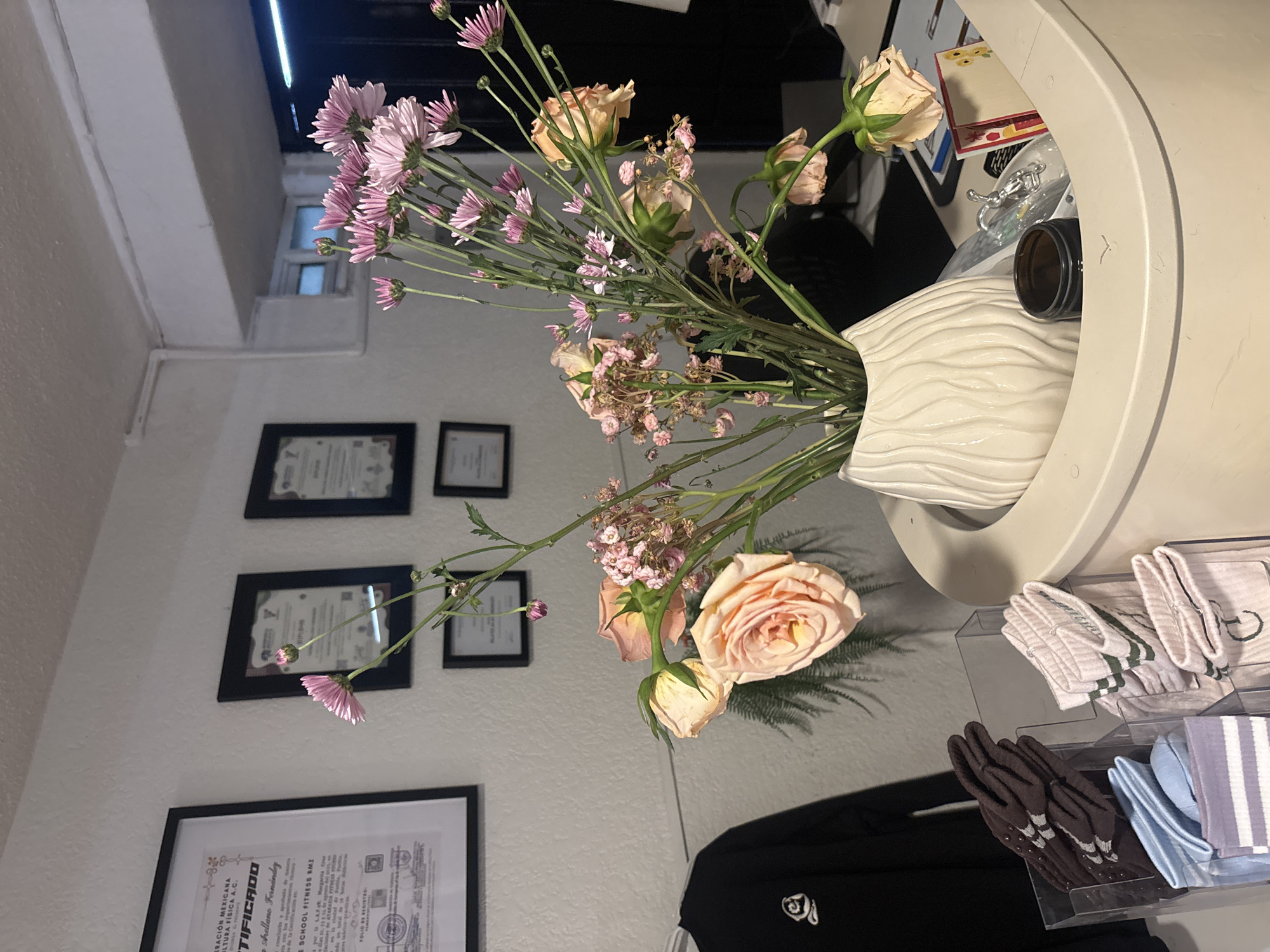 The image shows a vase of flowers on a counter. The flowers are a mix of pink and peach-colored varieties, with some buds and greenery. The vase is white with a textured surface. The counter is light-colored. Behind the flowers are several framed certificates hanging on a white wall. The wall appears to be in a fitness business. Below the counter, there are several drawers filled with folded socks. The bottom left corner of the image contains a portion of a framed certificate with the words "ERACIÓN MEXICANA," "ALTURA FÍSICA A.C.," "TIFICADO," and "Arellano Fernández" visible. The certificate also mentions "SCHOOL FITNESS SMZ." There is a black piece of clothing. Based on the visible information, the location of the picture is in Puebla, Mexico.