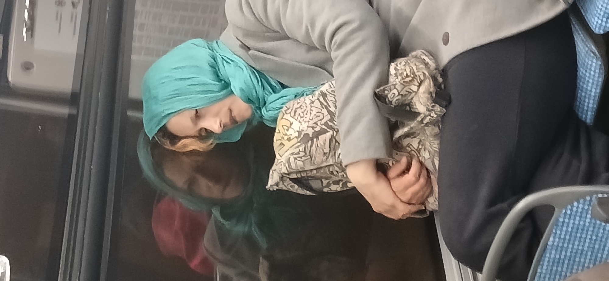 A woman is depicted resting or sleeping while seated in what appears to be a public transport vehicle in Moscow, Russia. She is wearing a teal-colored headscarf that covers her hair, a light grey long-sleeved jacket or cardigan, and dark trousers. Her eyes are closed, and her head is gently leaning against a dark, reflective surface, likely a window or a panel within the vehicle.
Her hands are clasped together in her lap, holding a piece of light-colored fabric with a dark, intricate pattern, which could be another scarf or a small bag. A sliver of light brown or blonde hair is visible near her temple, peeking out from under the headscarf.
The reflective surface next to her shows a clear, inverted reflection of her face and headscarf, along with a blurred reddish shape, possibly indicating ambient light or another element within the vehicle. To her right, part of a seat with a blue and white checkered or patterned upholstery is visible, suggesting she is on a bus or train. The background on the left consists of a dark panel with some indistinct grey elements, possibly part of the vehicle's interior structure or exterior view. The overall impression is one of peaceful rest during transit. No visible text is present.