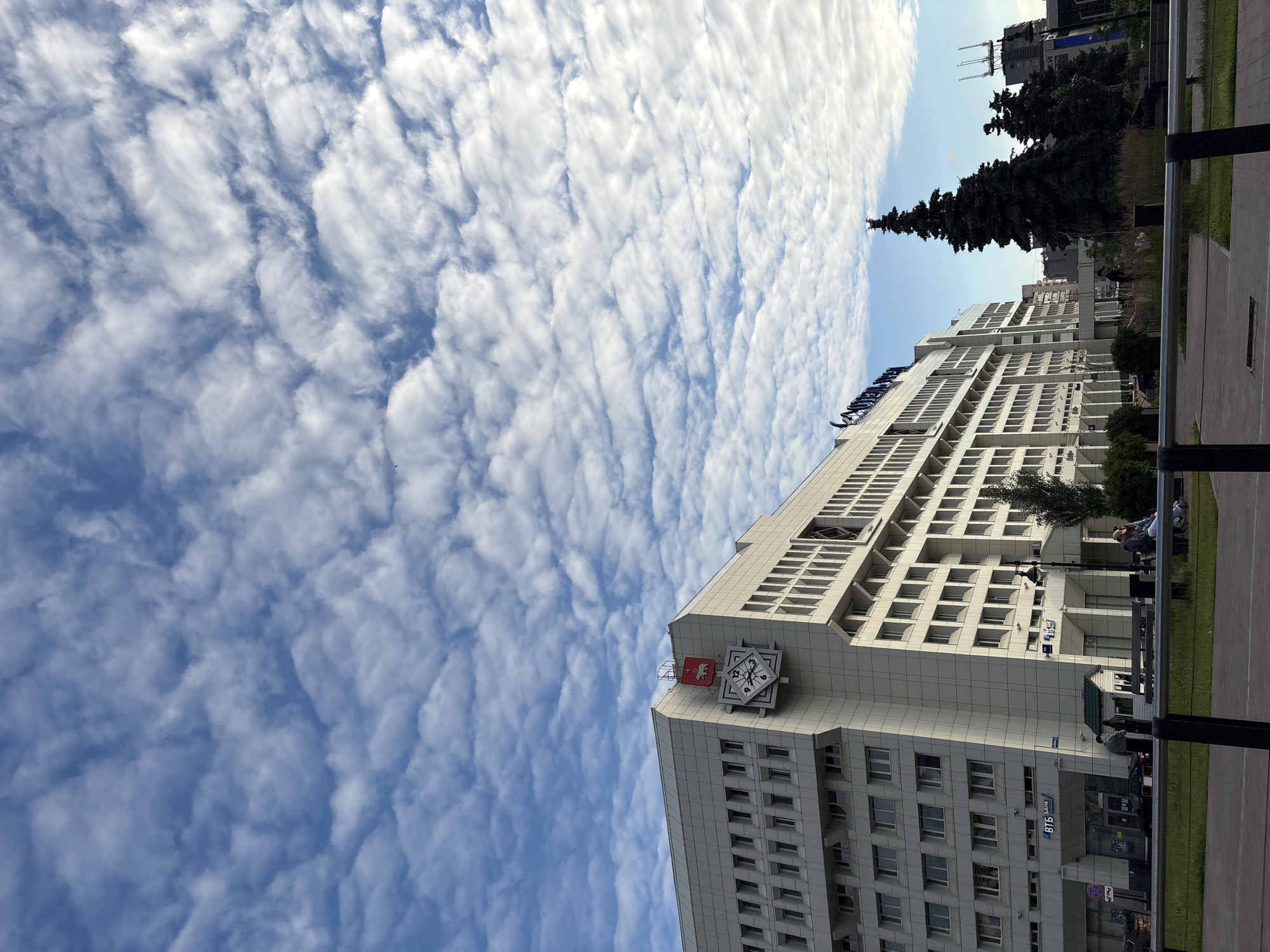 The image shows a wide view of a modern, multi-story white building with a distinctive architectural design, featuring a prominent clock on its facade. The building is located in the city of Perm, Russia. The sky above is a bright blue, filled with scattered, wispy white clouds. In front of the building, there is a sidewalk area with benches and some greenery, including several tall evergreen trees. A few people are visible, some walking and others sitting on the benches. The overall scene suggests an urban environment on a pleasant day.