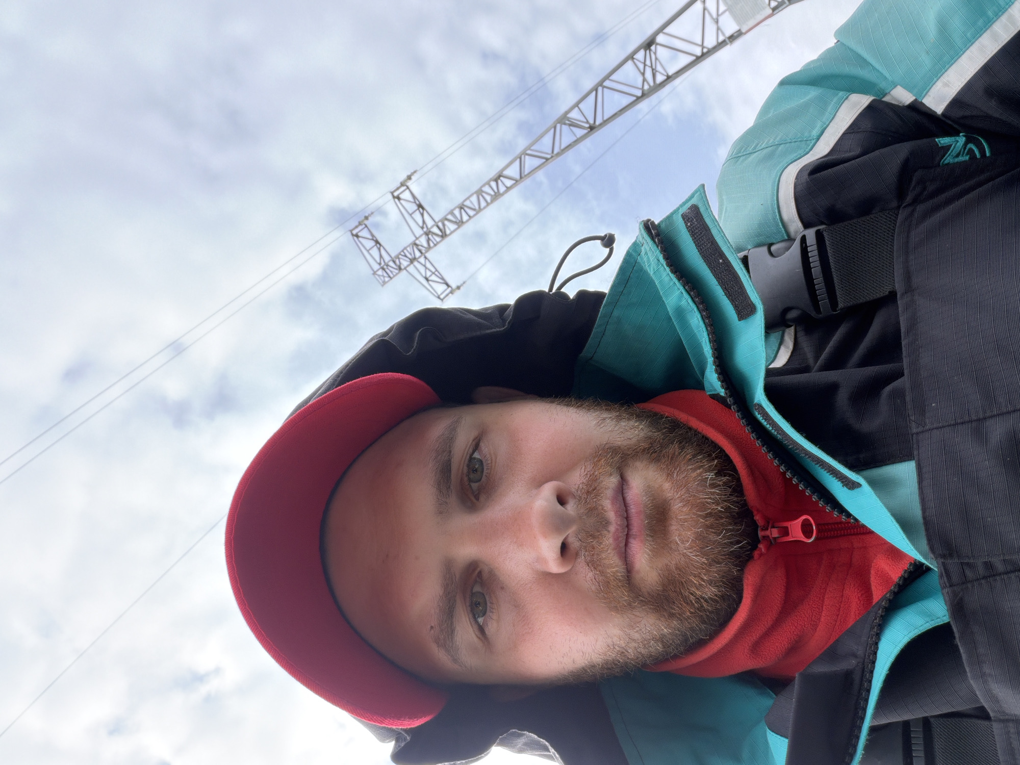 A man with a beard and a red baseball cap is seen from a low angle against a cloudy sky. He is wearing a red hoodie and a turquoise and black jacket, which is partially zipped. Above him is a metal structure, possibly part of an electrical tower. Without further context, I can't confirm the location to be Chaikovsky, Russia or identify any actions, interactions, or subjects besides the man and the tower.