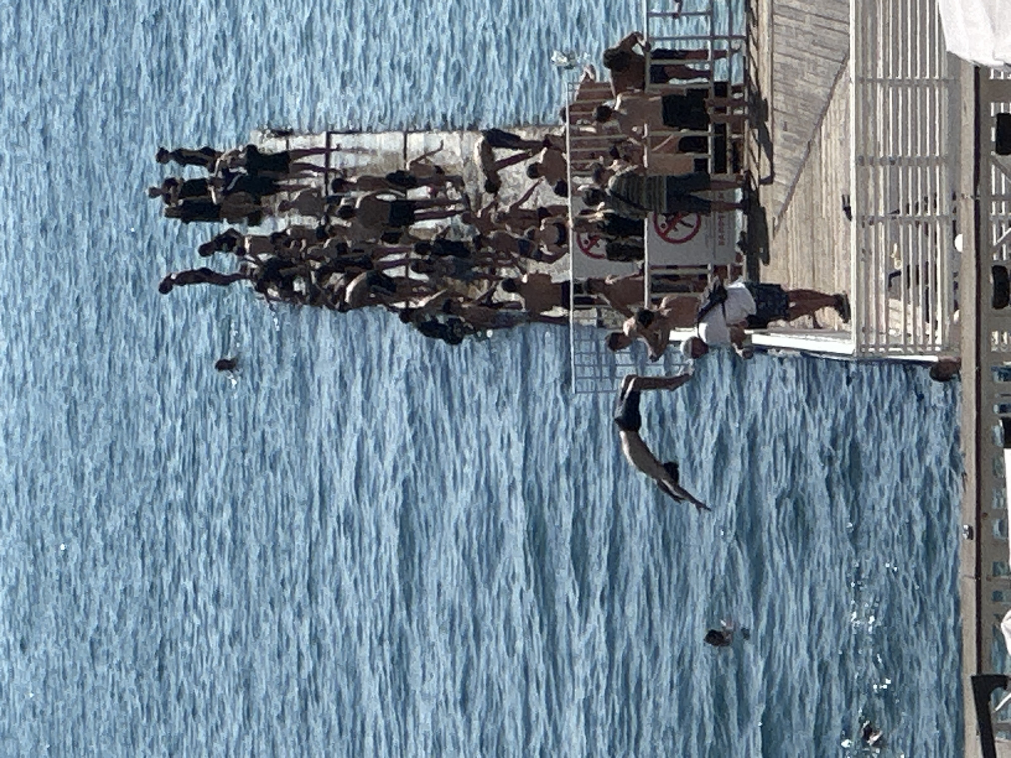 This image depicts a crowded wooden pier on the Black Sea coast in Sochi, Russia. Numerous people are present on the pier, with many standing and some in the water. One individual is captured mid-air, performing a dive from the pier into the water. The sea is a deep blue, with visible waves. The pier is constructed from wood and has railings. There are signs on the pier, though their text is largely illegible. The overall scene suggests a popular recreational area during a warm day.