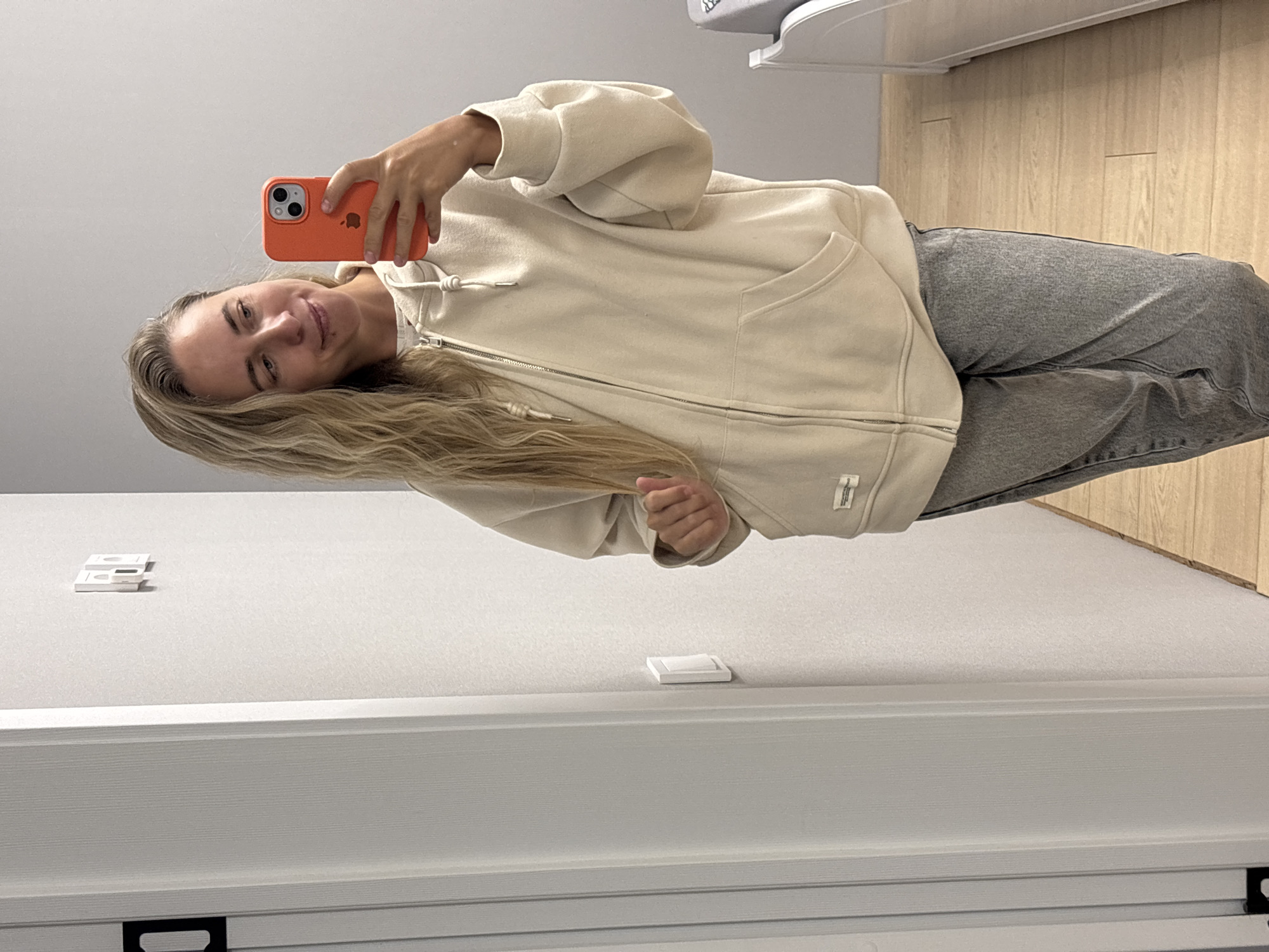 A woman with long blonde wavy hair is standing in front of a mirror, taking a selfie. She is wearing a beige zip-up hoodie and grey jeans. The hoodie has a white drawstring and a small tag on the left side. She is holding an orange iPhone in her right hand, which is partially visible in the frame. The background appears to be an indoor setting, possibly a room with light grey walls and a white door on the left. A light switch is visible on the wall. The floor is light wood. There is a hint of a white bed frame or piece of furniture on the right side of the image. The lighting suggests it is daytime. The image does not provide enough information to determine the exact location within the city of Chaikovsky, Russia.