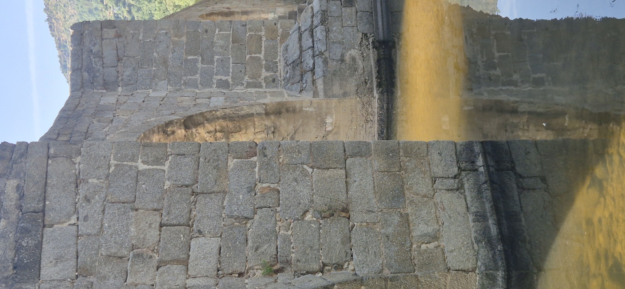 The image shows a close-up view of a stone bridge and its reflection in the water below. The bridge is constructed from large, rough-hewn granite blocks, with visible mortar lines between them. The stonework appears weathered and aged. On the left side of the image, a thick, vertical section of the bridge wall is prominent, showing rows of carefully stacked stones. To the right, an arched opening is visible within the bridge structure, with sunlight illuminating the interior, creating a warm glow. Further to the right, another section of the bridge wall and a smaller archway are visible. The water is shallow and has a yellowish-brown hue, likely due to sediment or the reflection of the surrounding environment. The reflection of the bridge is clearly visible in the water, with some distortion due to the ripples. In the background, a hillside covered in trees and vegetation can be seen under a bright blue sky. The overall scene depicts a historic stone bridge in a natural, outdoor setting, likely near a body of water.