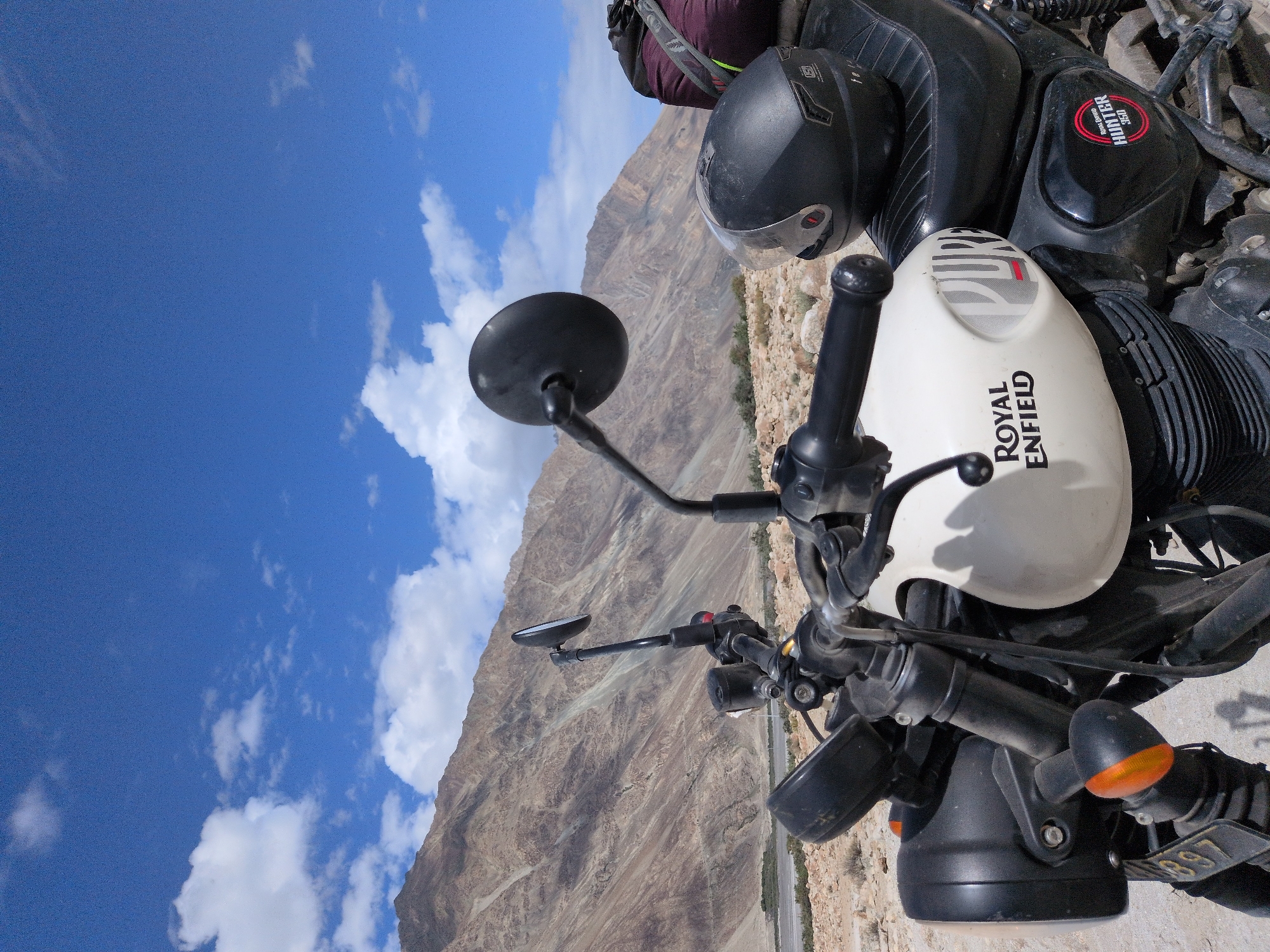 A motorcycle, identified by its "Royal Enfield HUNTER 350" badging, is parked on a rocky, arid terrain under a bright blue sky with scattered clouds. The motorcycle, primarily white and black, is positioned in the foreground, angled towards the viewer's right. It features a prominent headlight, handlebars with mirrors, a fuel tank with "ROYAL ENFIELD" and "PURE" branding, and a black leather seat. A black helmet rests on the seat. In the background, rugged, light-brown mountains rise steeply, their slopes marked with various textures and shadows. A dry riverbed or valley can be seen at the base of the mountains, with sparse vegetation. The overall impression is a solitary scene in a remote, mountainous landscape, likely within the region of Sasoma, India, given the context provided. No people are visible in the image.