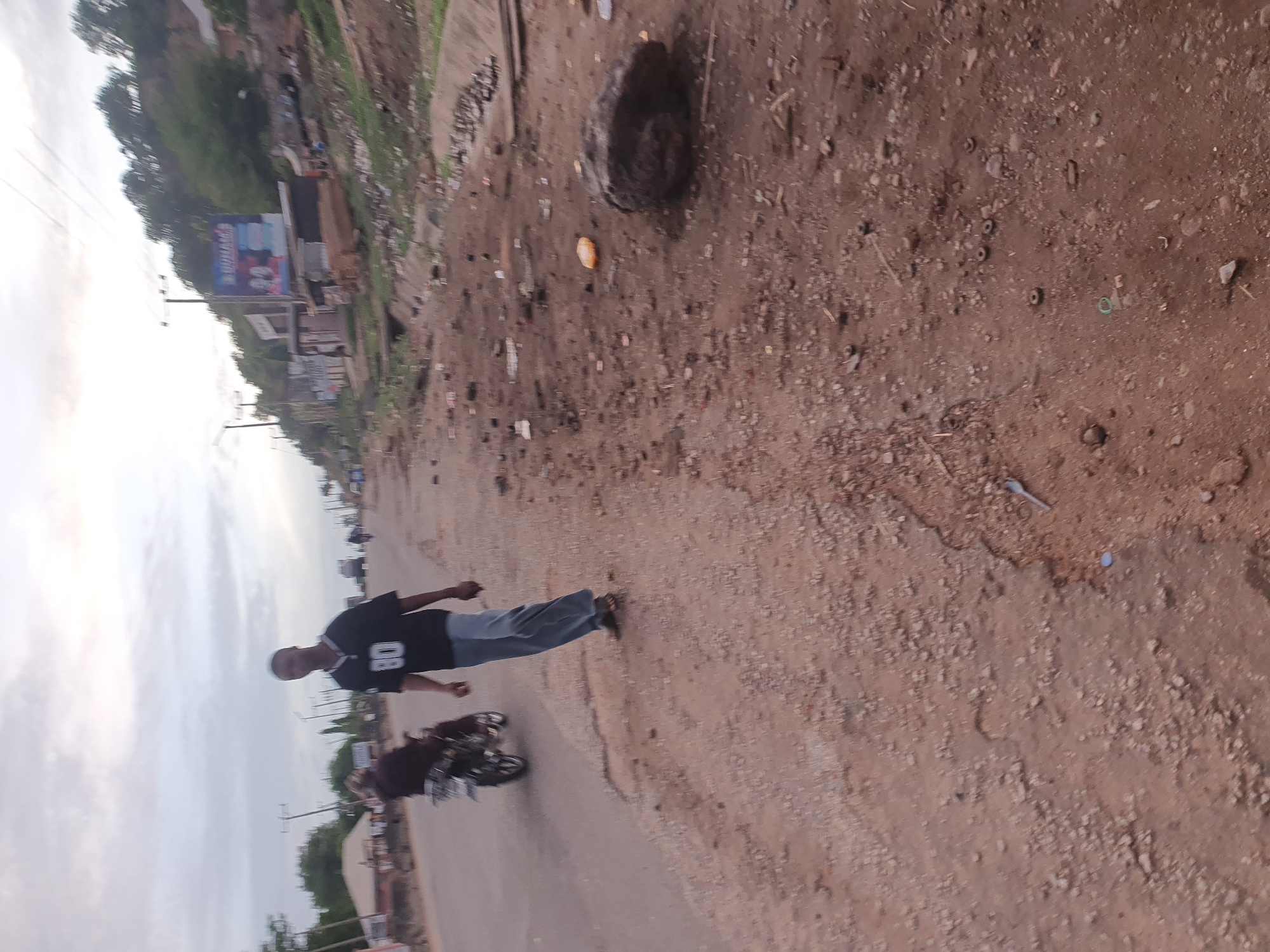 The image shows a street in Lokoja, Nigeria. A man wearing a black jersey with the number 08 and grey pants is walking along the side of the road. A motorcycle is parked nearby. The road is unpaved and dusty with debris. In the background, there are buildings, trees, and utility poles. A billboard is visible on one of the buildings.