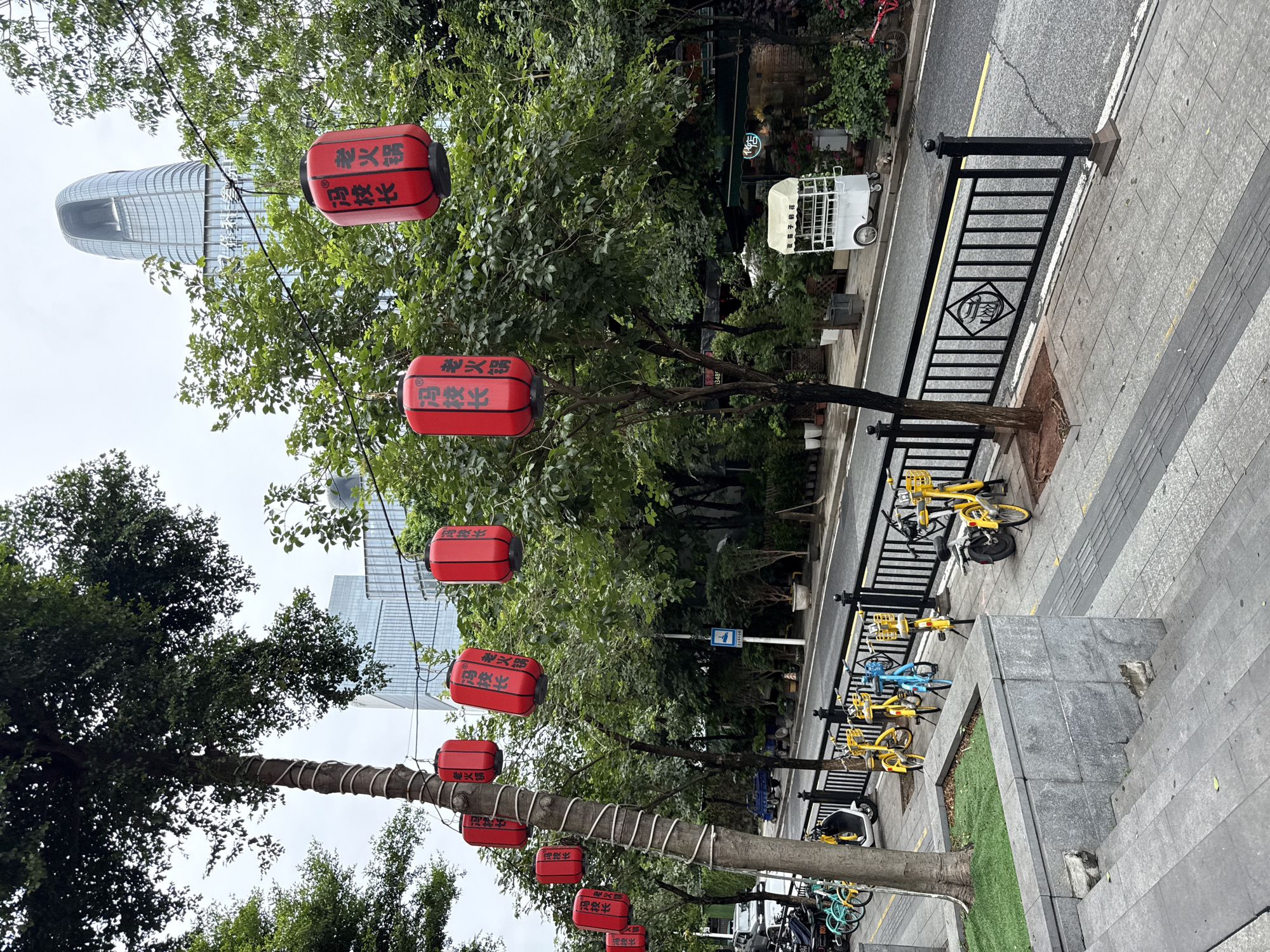 The image depicts a street scene in Haizhu District, China, on an overcast day. Red paper lanterns with Chinese characters are strung across the street, hanging from wires and trees. The trees lining the street are lush with green foliage. In the background, a modern skyscraper with a unique architectural design is visible.
The street itself is paved and has a yellow line marking the lane. A black metal fence runs along the sidewalk, which is made of grey paving stones. Several yellow and blue shared bicycles are parked along the sidewalk behind the fence. A white cart with a canopy, possibly a vendor's stall, is positioned further down the street. The ground appears wet in places, suggesting recent rain. There are no people clearly visible in the foreground of the image. The overall atmosphere is that of a bustling urban environment with cultural elements integrated into the cityscape.