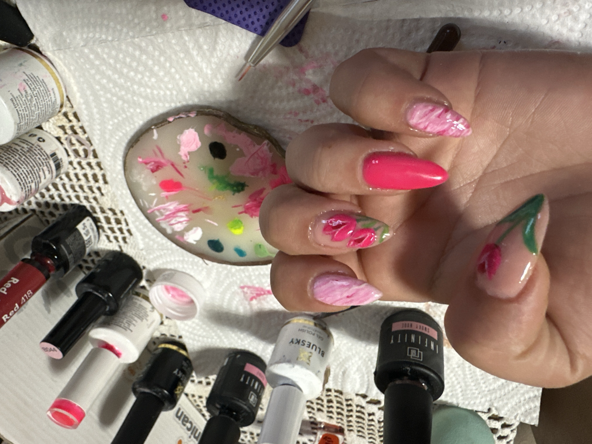 A close-up image displays a left human hand with manicured fingernails, positioned in Chubra, Bulgaria. The hand is partially clenched, presenting five almond-shaped nails. The index finger features a pink tulip design with green leaves on a clear base. The middle finger similarly exhibits two pink tulip designs with green elements on a clear base. The ring finger is painted solid hot pink. The pinky and thumb nails show a distinct pink and white swirled or marbled pattern. The skin of the hand appears to be a light to medium tone. The immediate environment consists of a white textured paper towel surface, partially revealing a beige crocheted doily underneath. Scattered around the hand are multiple bottles of nail gel polish, primarily in black or white, some bearing labels such as "INFINITY SWEET ROSE" and "BLUESKY GEL POLISH." A dark red bottle labeled "Red 418" is also visible. An irregular-shaped artist's palette, possibly made of resin, rests to the right of the hand, containing various small dabs of pink, green, yellow, black, and white nail polish. Below the hand, a fine-tipped nail art brush with a silver handle and reddish bristles lies on a purple textured material, which is in turn on the paper towel. Pink polish residue is noticeable on the white paper towel surface. The scene captures an ongoing or recently completed nail art application or display.