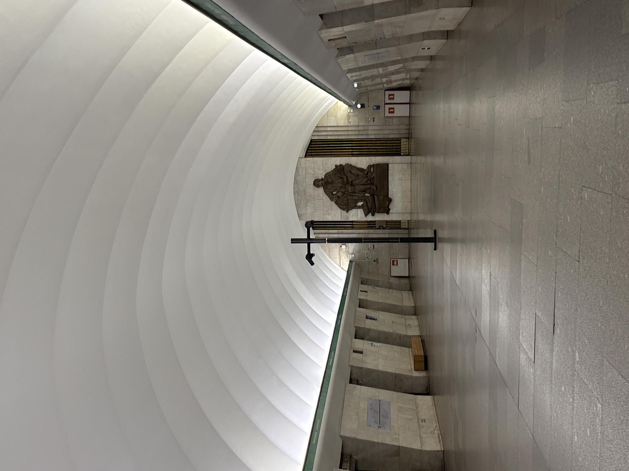 This image displays the empty platform of Vasileostrovskaya metro station in Saint Petersburg, Russia. The station features a high, white, vaulted ceiling constructed from numerous distinct, smooth, arched segments. This ceiling is illuminated by continuous strip lighting recessed along the upper perimeter of both platform walls.
The platform walls are composed of light-colored stone or concrete, forming a repeating pattern of projecting, angled, trapezoidal pillar-like structures. On the left wall, a single wooden bench is visible.
At the far end of the platform, a large, dark bronze bas-relief sculpture is mounted centrally. The sculpture depicts a seated male figure, identifiable as Mikhail Lomonosov, accompanied by a globe and several books. This sculpture is set against a textured wall composed of vertical dark metallic slats. To the right of the sculpture, two white doors with prominent red square markers are visible.
A singular black, T-shaped pole stands upright in the center of the gray-tiled platform floor. The floor exhibits subtle variations in color and texture across its rectangular tiles. Indistinct signage is affixed to some of the angled wall segments, and a red-and-white rectangular sign is positioned on the right wall. No individuals are present within the scene.